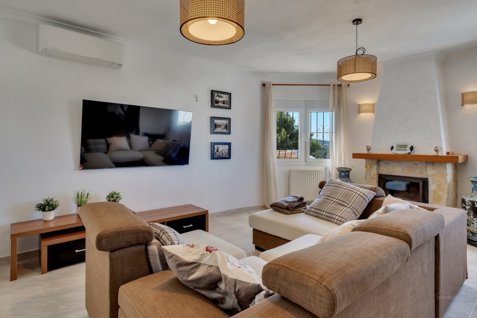Lichte woonkamer met natuurstenen open haard, airconditioning en wandgemonteerde tv. Voorzien van rieten hanglampen, radiatorverwarming en een ruime modulaire zithoek.