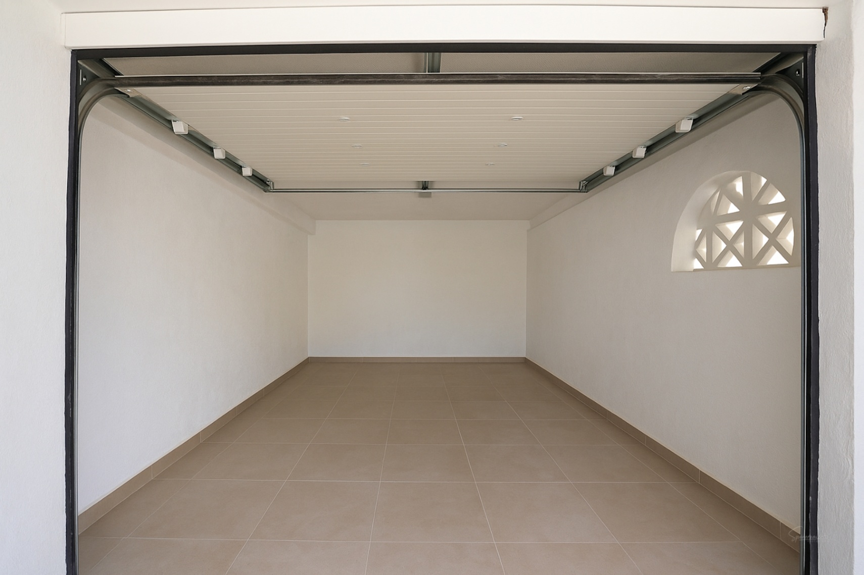 Privégarage met tegelvloer, witte muren en een automatische sectionaaldeur. Een decoratief boograam met rasterwerk zorgt voor natuurlijk licht.