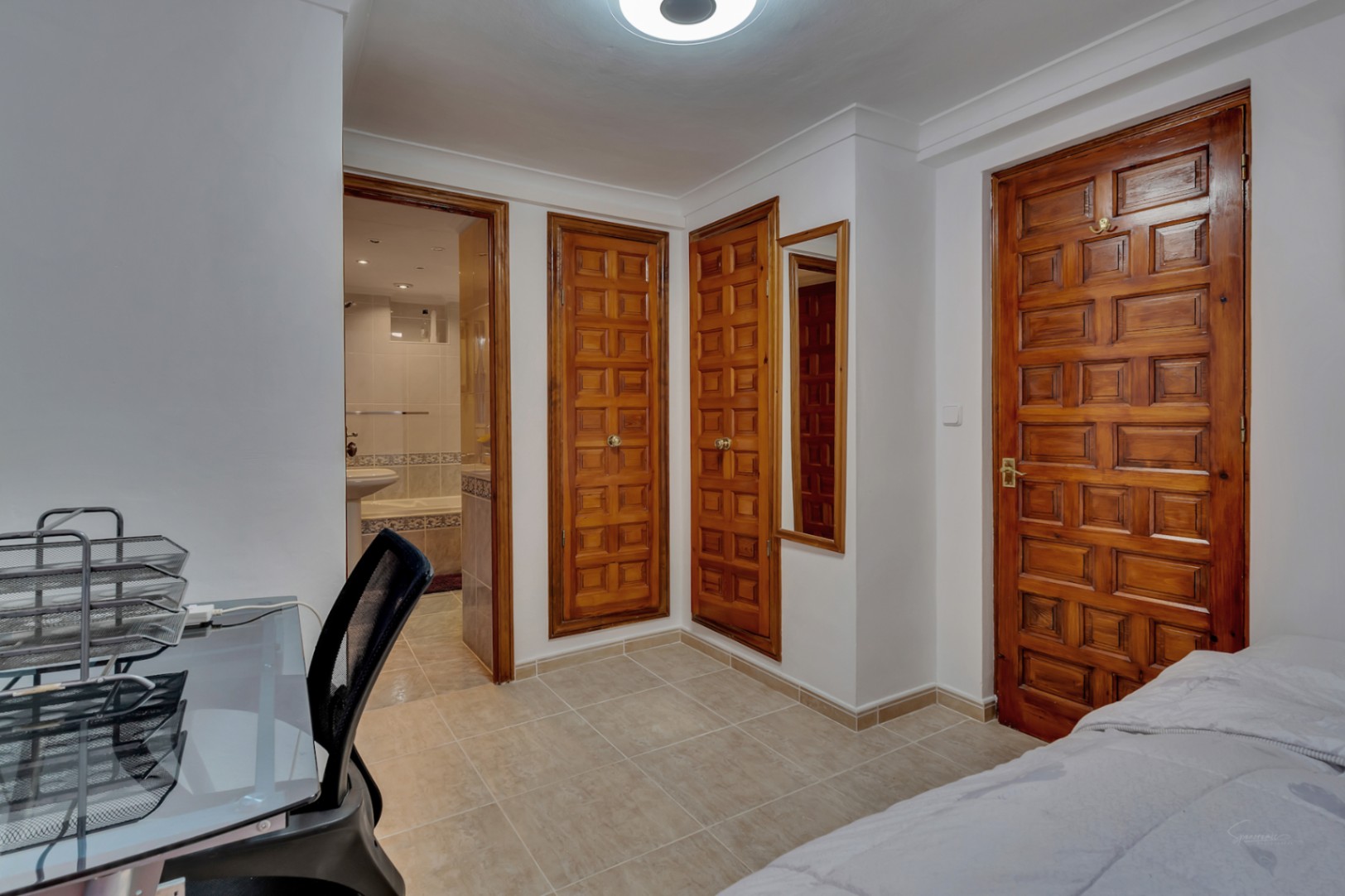 Slaapkamer met ingebouwde kasten van massief hout, keramische tegelvloer en directe toegang tot een en-suite badkamer. Voorzien van een functionele werkplek.