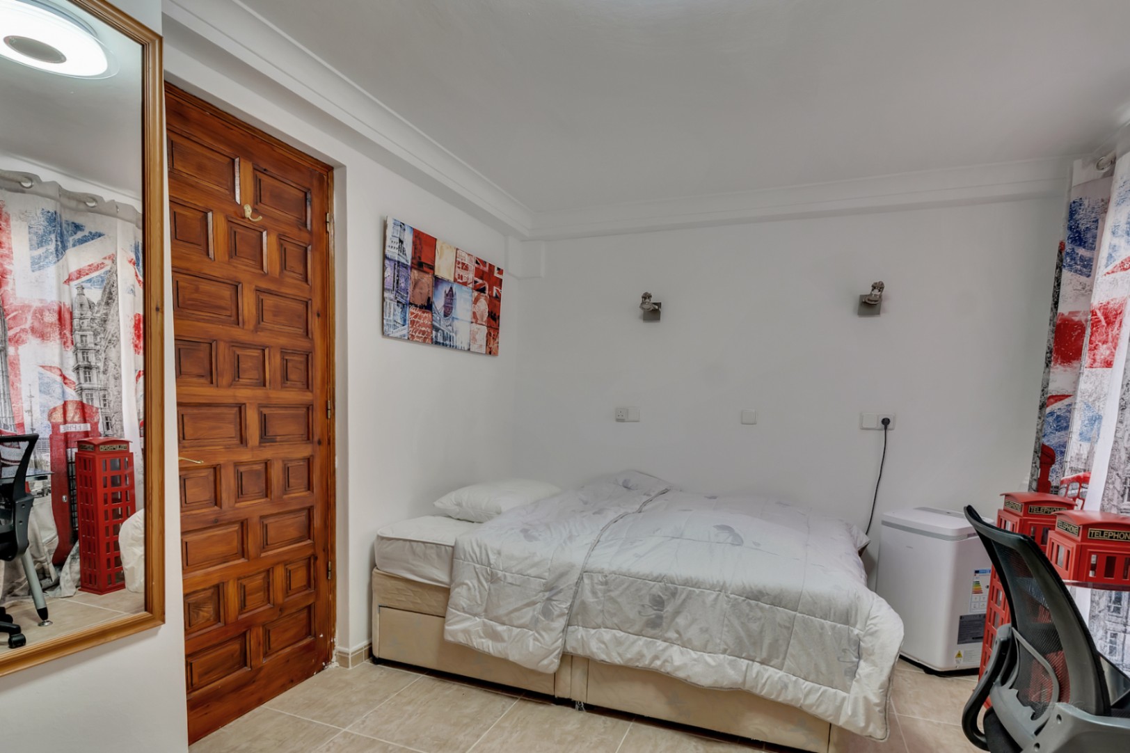 Slaapkamer met keramische tegelvloer, een massief houten paneeldeur en wandverlichting. De ruimte is voorzien van een grote spiegel en unieke thematische decoratie.