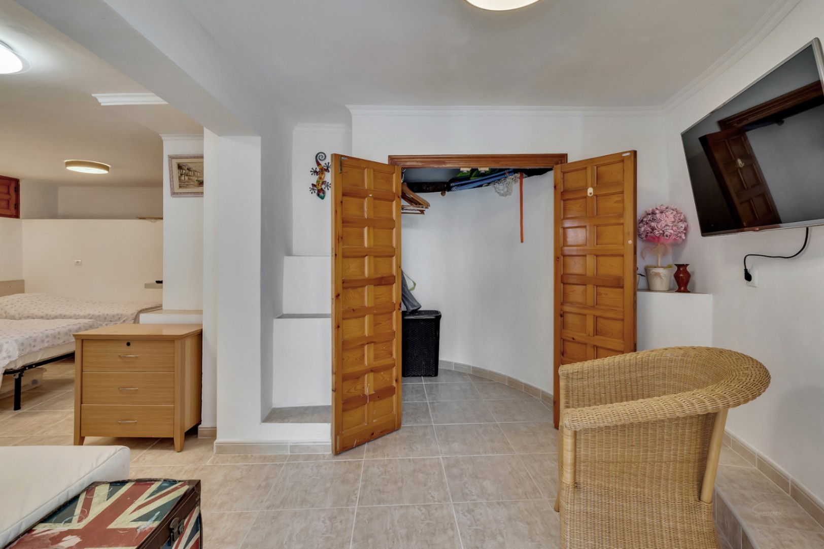 Slaapkamer op de benedenverdieping met tegelvloer, houten inbouwkast en een zithoek voorzien van een televisie en rotan fauteuil.