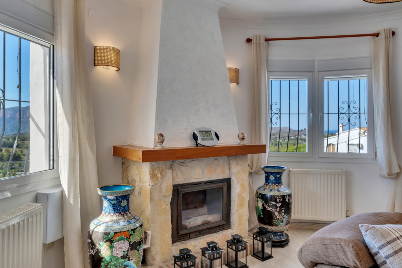 Woonkamer met een natuurstenen open haard en houten plank, geflankeerd door ramen met uitzicht op de Montgó en de zee, voorzien van radiatoren en wandverlichting.