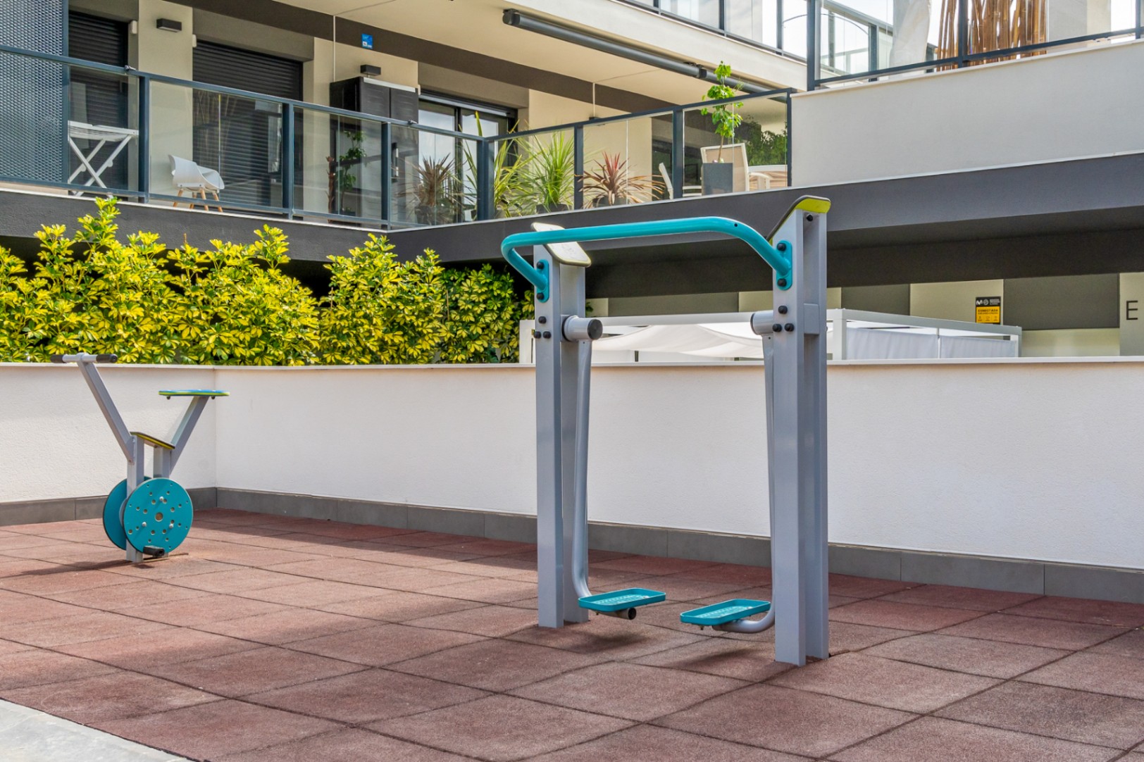 Buitenfitnessruimte voorzien van moderne trainingsapparatuur op een rubberen veiligheidsvloer, gelegen nabij de groene zones van het moderne wooncomplex.