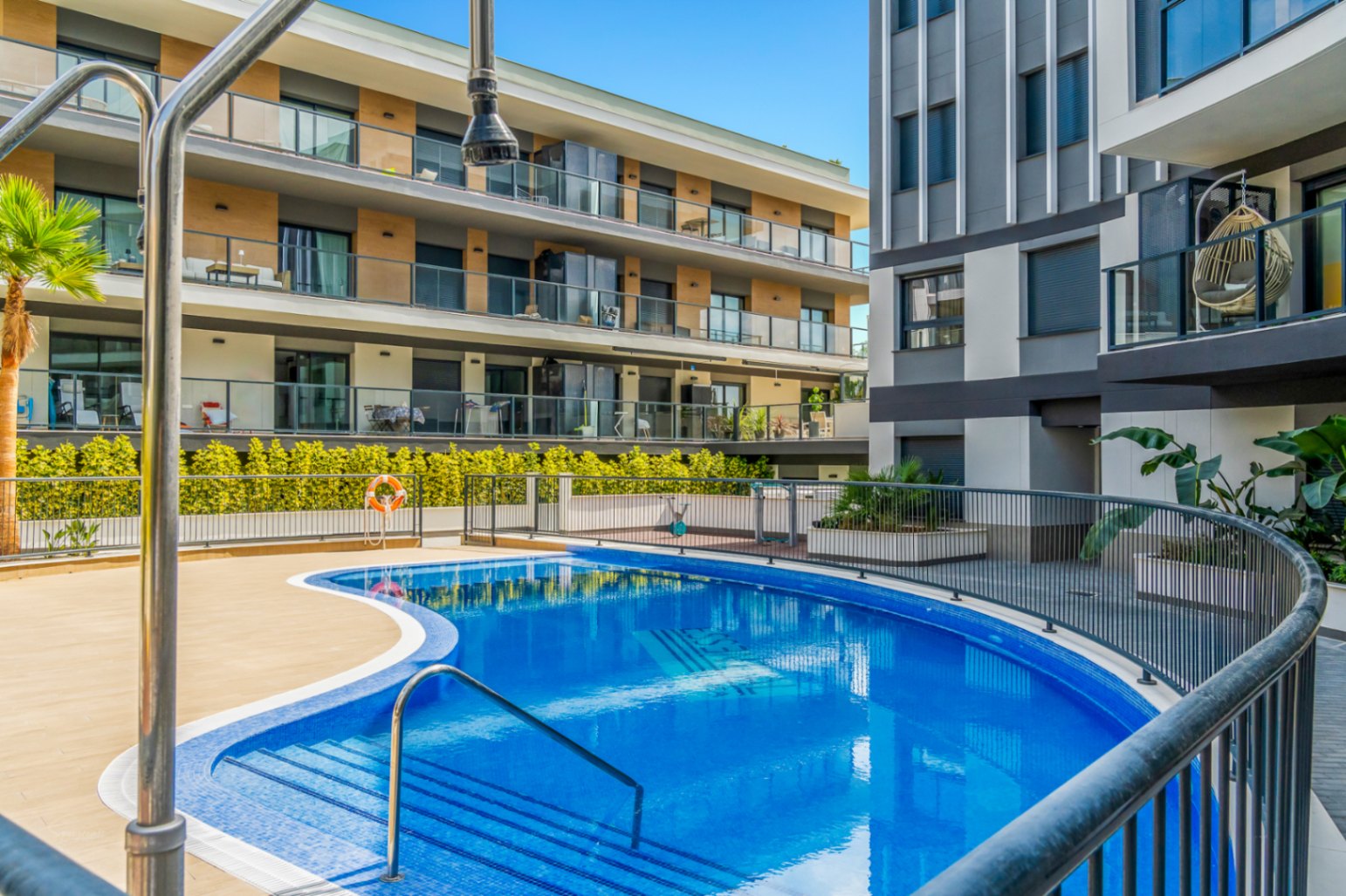 Gemeenschappelijk zwembad met ronde vormen, omgeven door een betegeld zonneterras en moderne appartementen met glazen balustrades en houten accenten.
