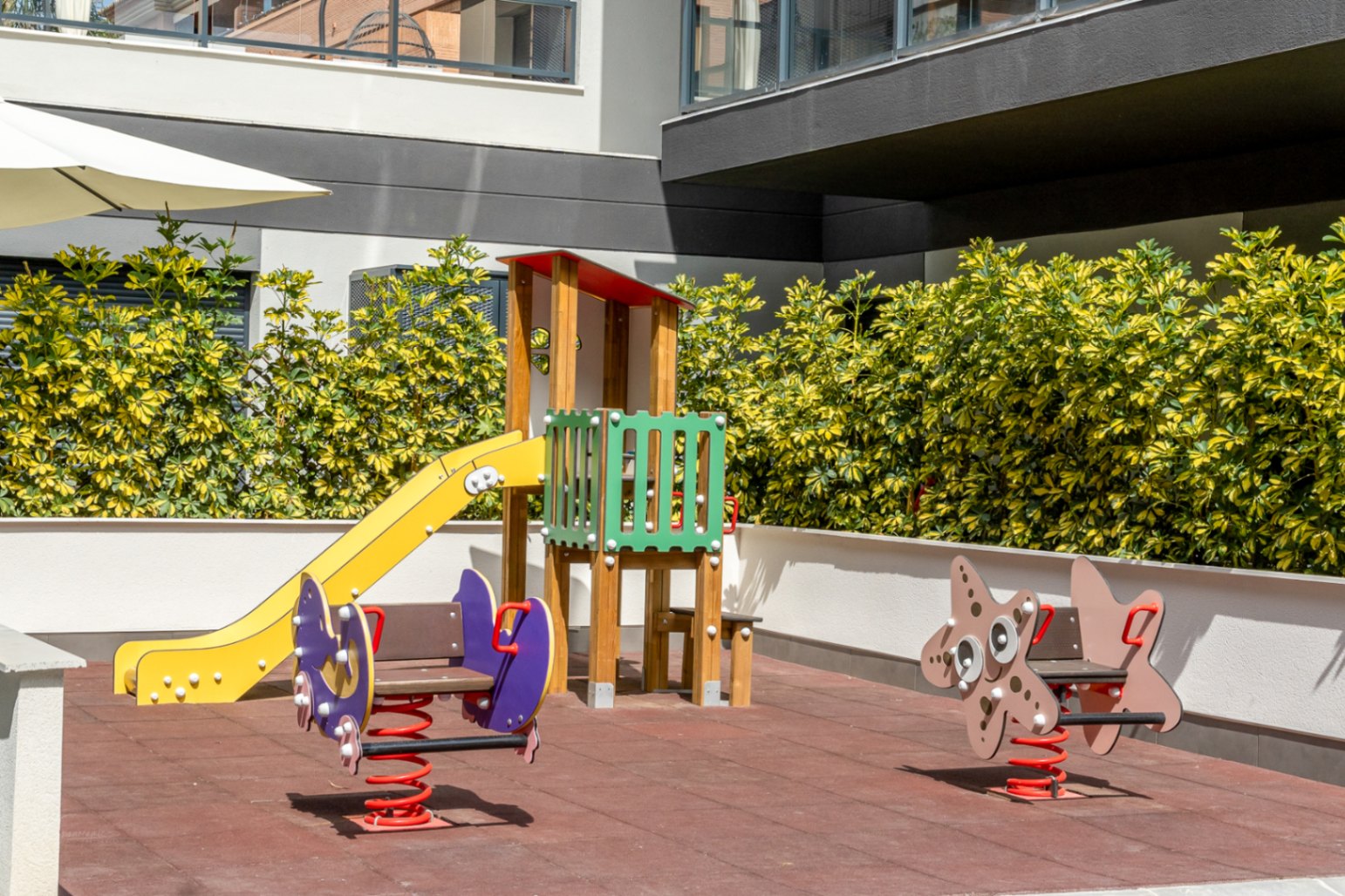 Gemeenschappelijke kinderspeelplaats met rubberen veiligheidsvloer, gele glijbaan en veertoestellen, omgeven door verzorgde hagen en moderne architectuur.