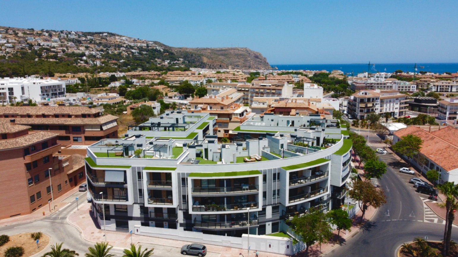 Luchtfoto van het moderne wooncomplex met gebogen witte architectuur, privédakterrassen met kunstgras en uitzicht op de Middellandse Zee in Jávea.