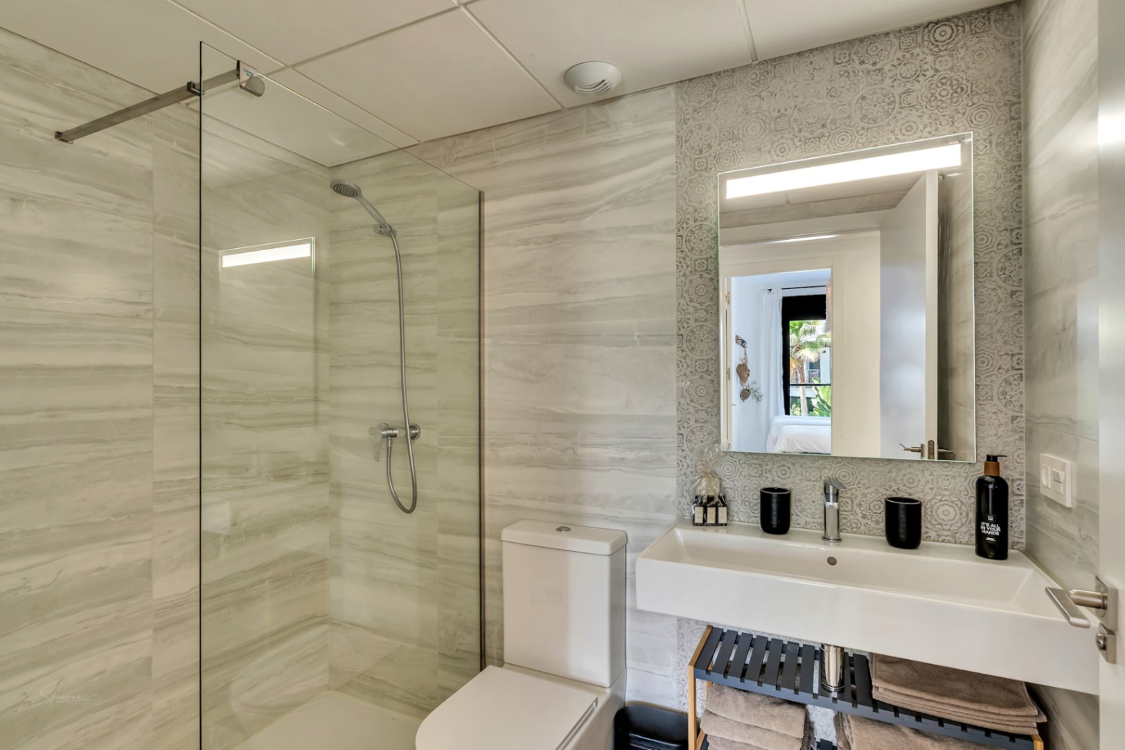 Moderne badkamer met inloopdouche, glazen wand en tegels met marmerlook. Voorzien van een zwevende wastafel tegen een gedecoreerde wand en een spiegel met geïntegreerde verlichting.