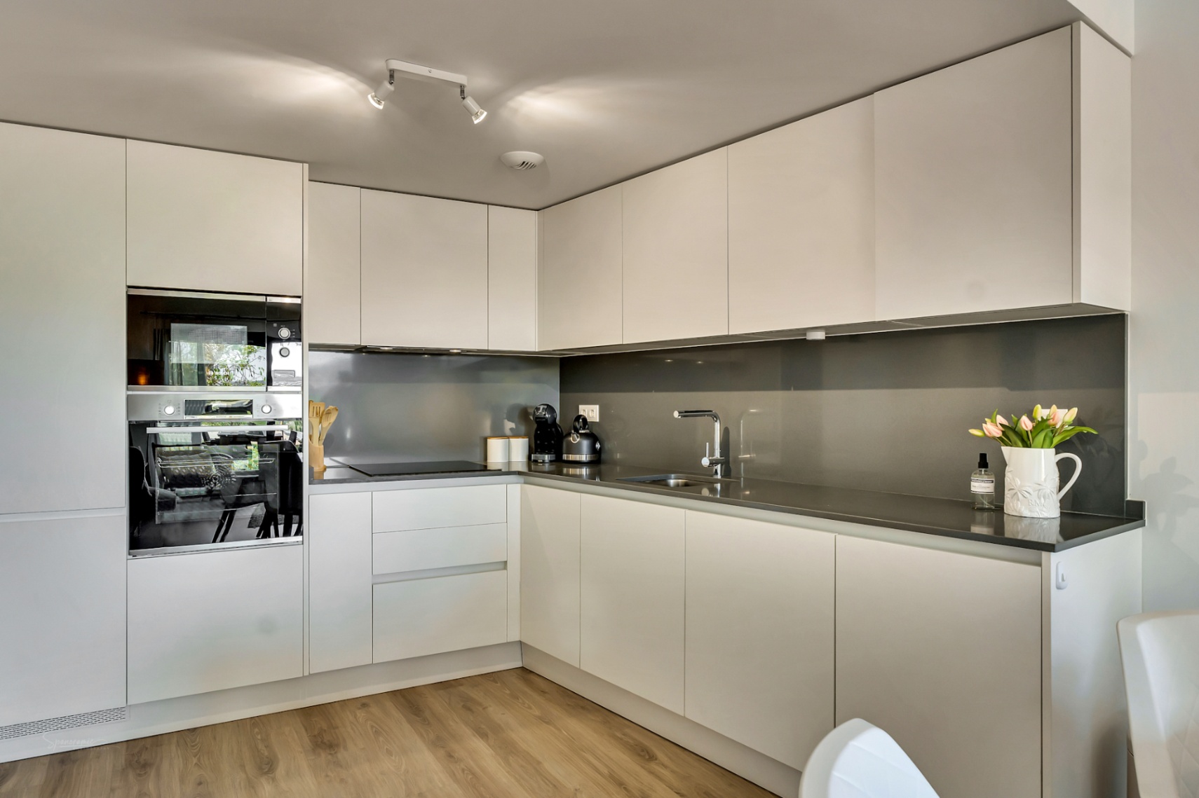 Moderne L-vormige keuken met greeploze witte kasten, een donkergrijs werkblad en geïntegreerde apparatuur. De houtlook vloer zorgt voor een warme, tijdloze uitstraling.