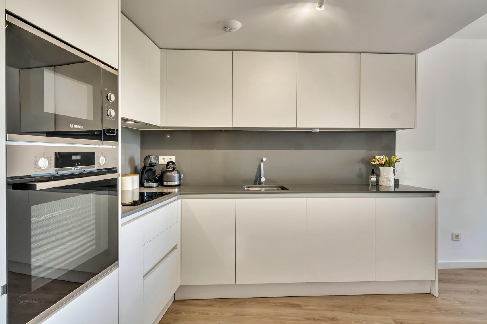 Moderne minimalistische keuken met greeploze witte kasten, een grijs werkblad en geïntegreerde Bosch-apparatuur op een lichte houten vloer.