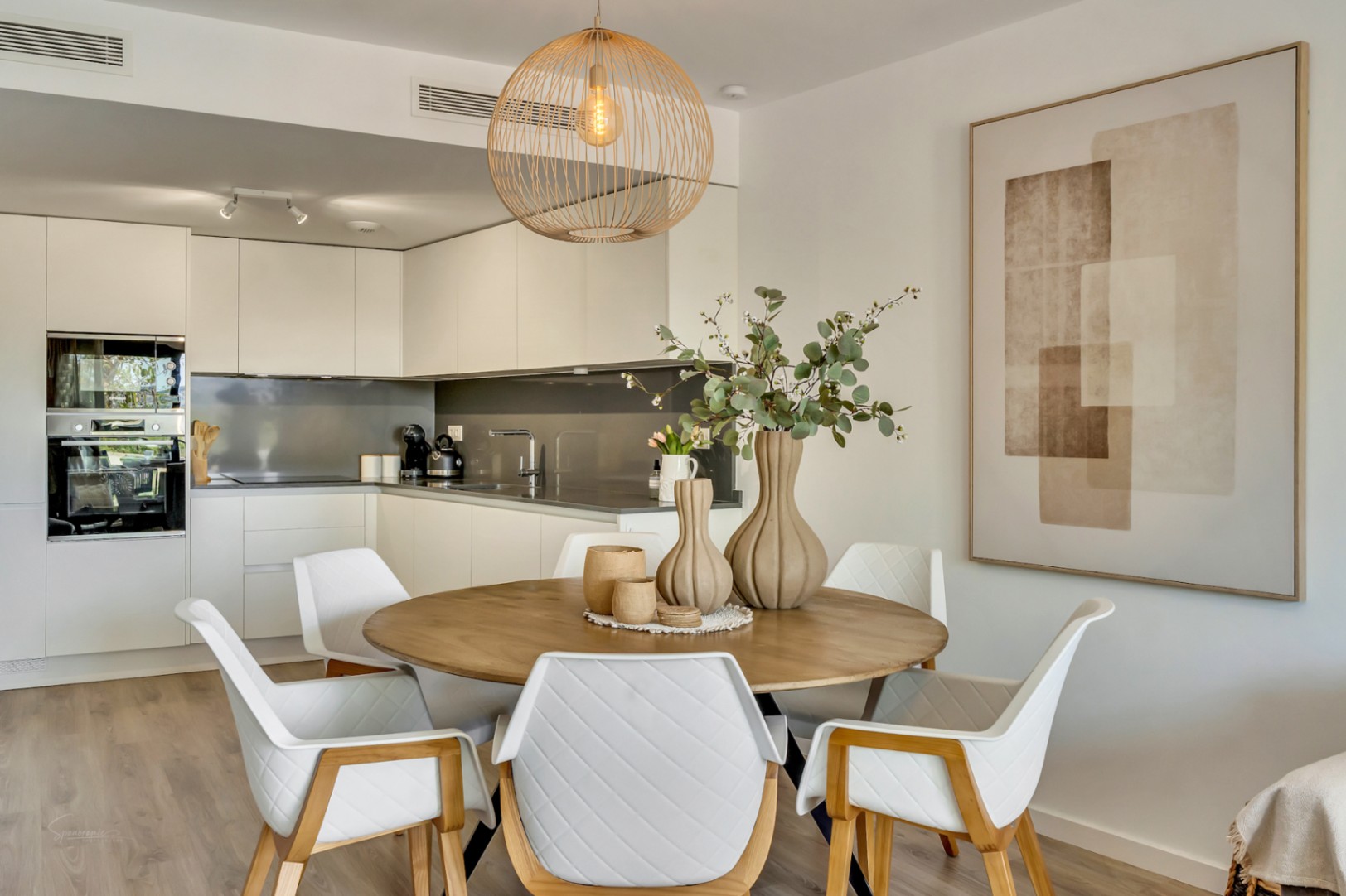 Open eetkamer met een ronde houten tafel, gewatteerde stoelen en een rotan hanglamp, vloeiend verbonden met een moderne witte keuken met inbouwapparatuur.