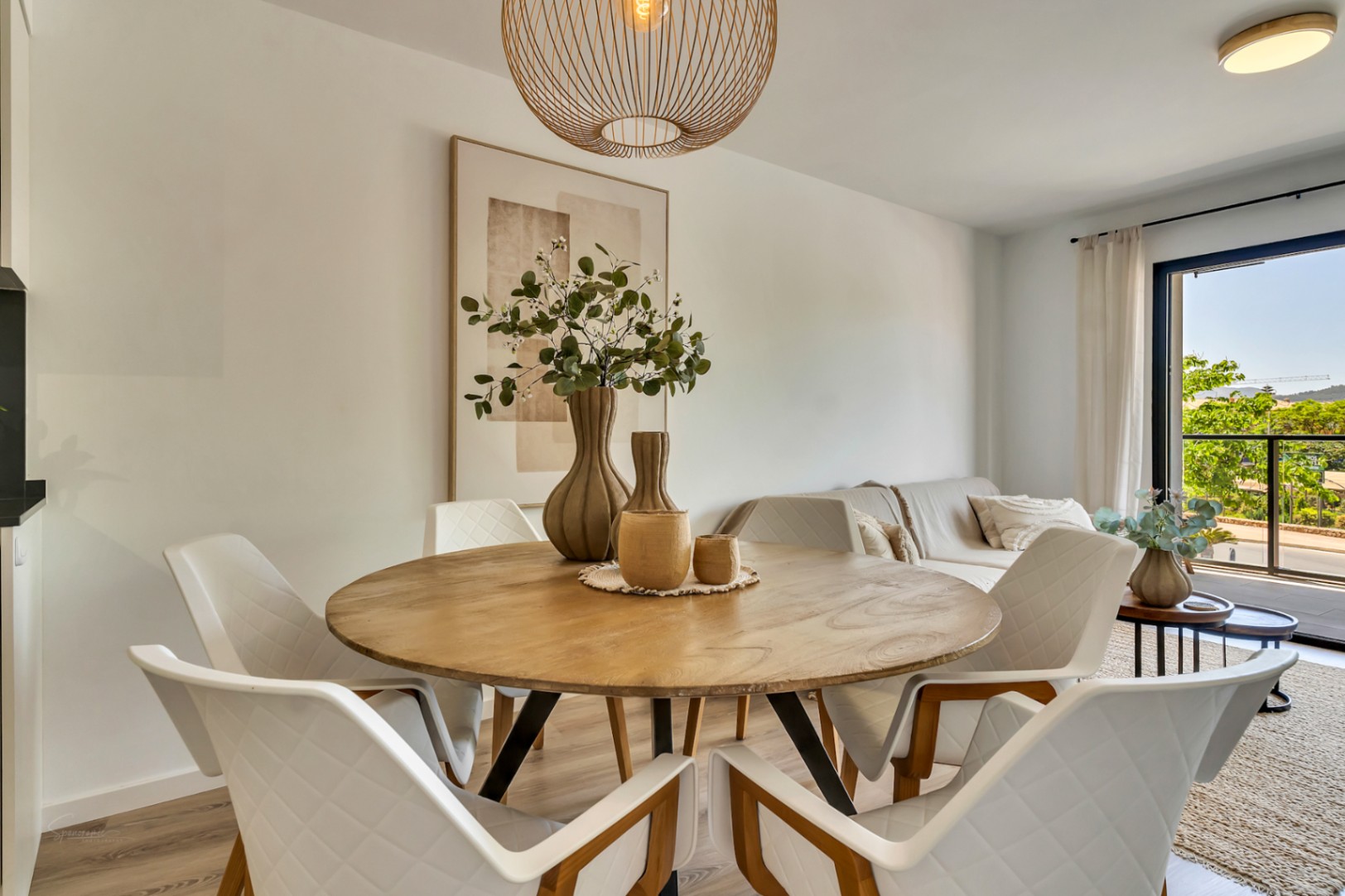 Open eetkamer met een ronde houten tafel, witte gecapitonneerde stoelen en een rotan hanglamp, verbonden met een lichte woonkamer en toegang tot het balkon.