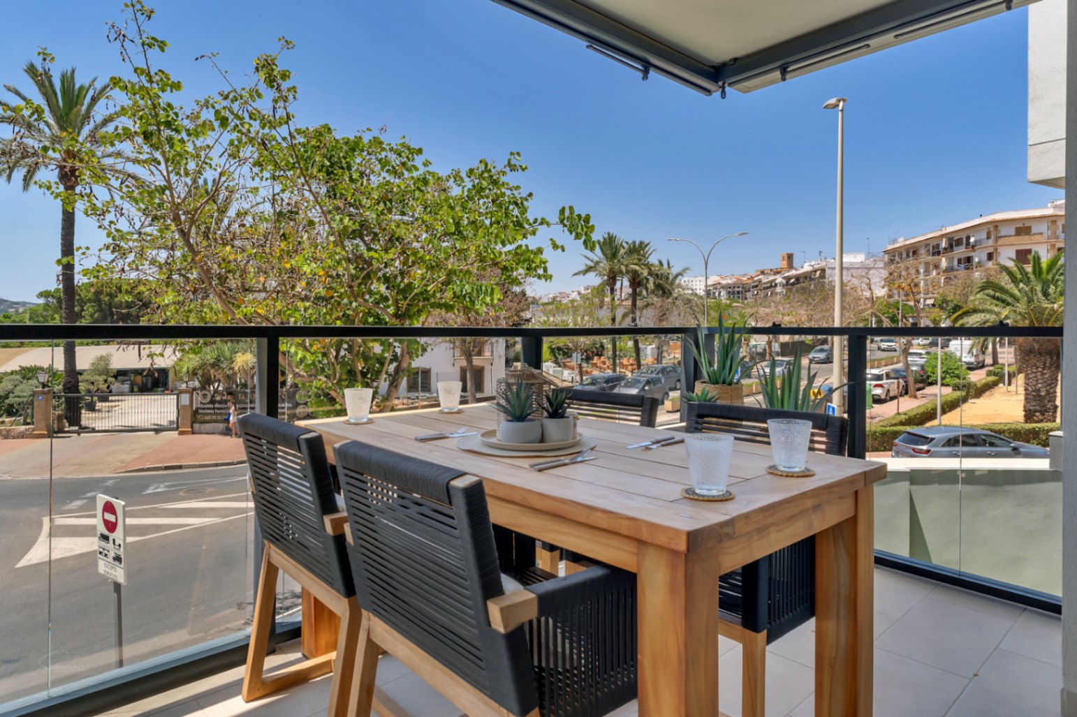 Privéterras met een houten eettafel, gevlochten stoelen en glazen balustrades met vrij uitzicht over de stedelijke omgeving en mediterrane beplanting.
