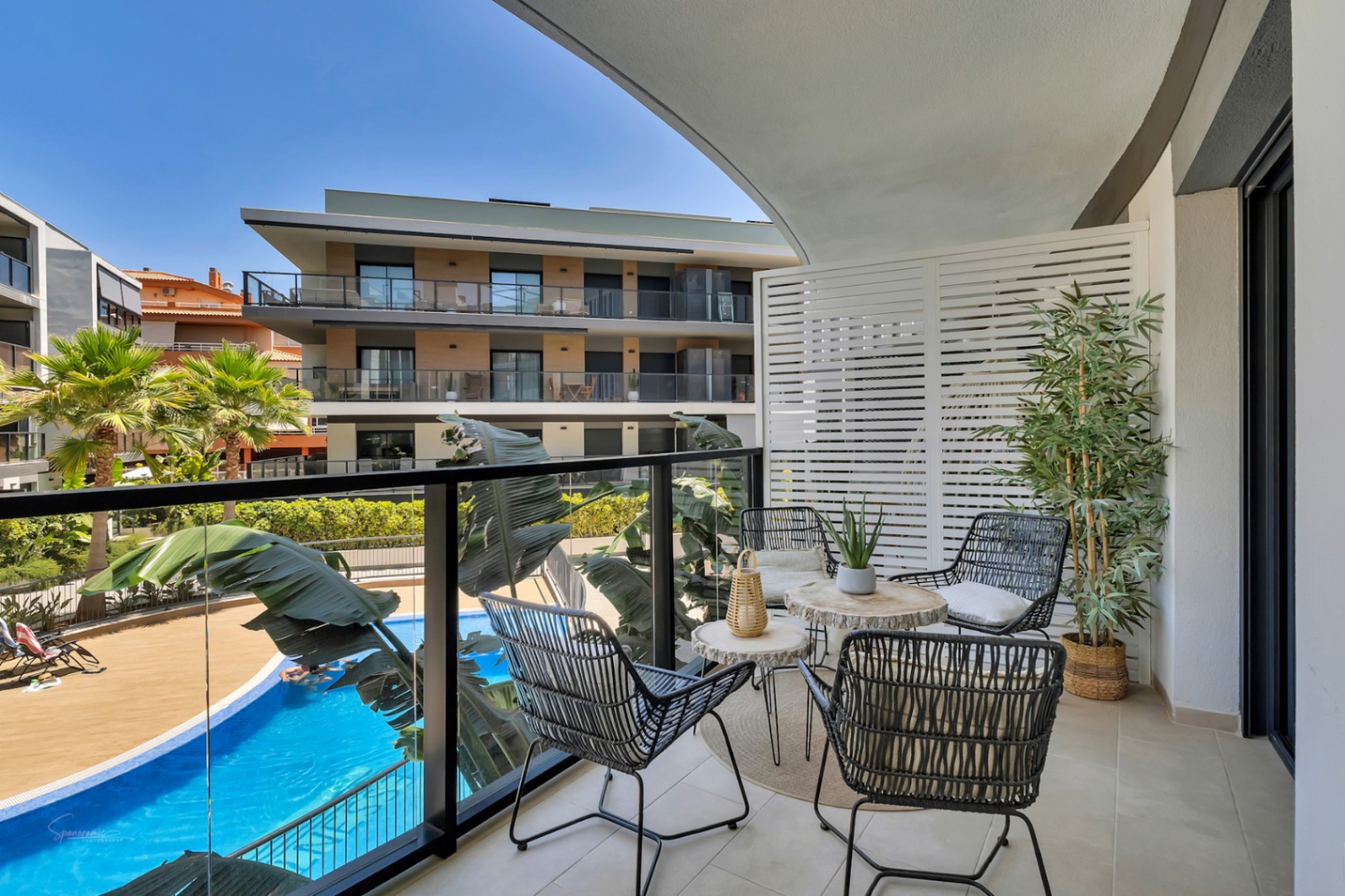 Privéterras met glazen balustrade, modern tuinmeubilair en uitzicht op het gemeenschappelijke zwembad en de aangelegde tuinen van het complex.