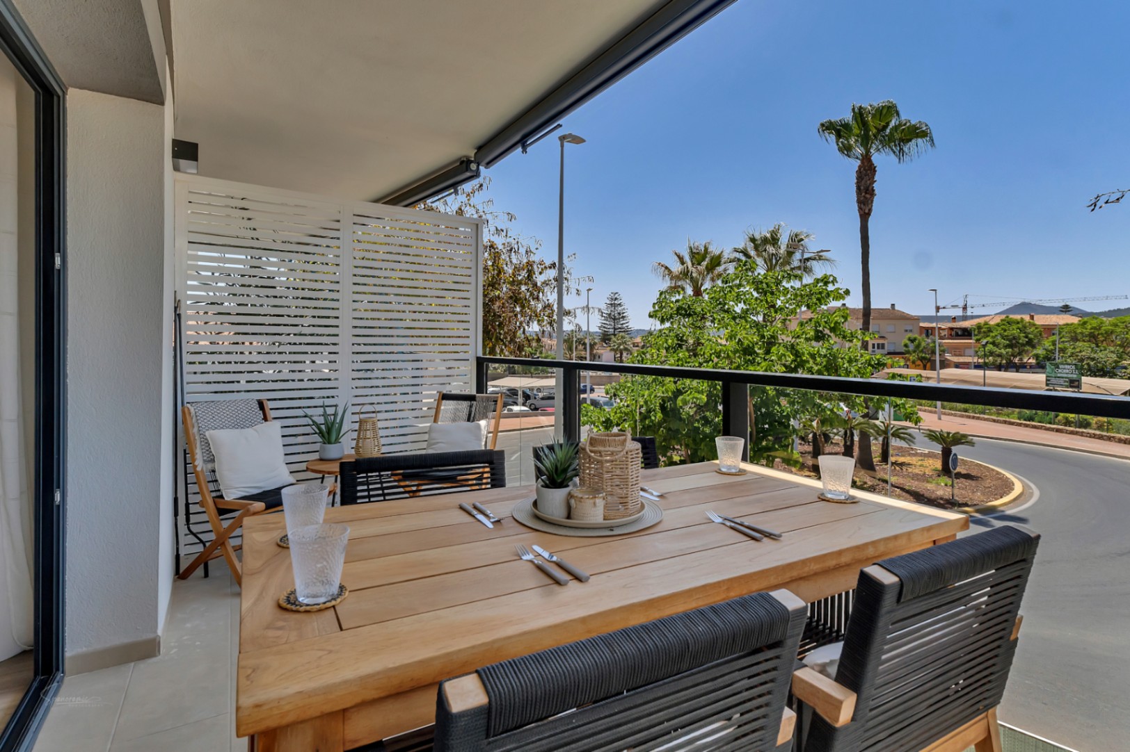 Privéterras met houten eettafel, gevlochten stoelen en glazen balustrade met vrij uitzicht op palmbomen en de omgeving van Jávea.