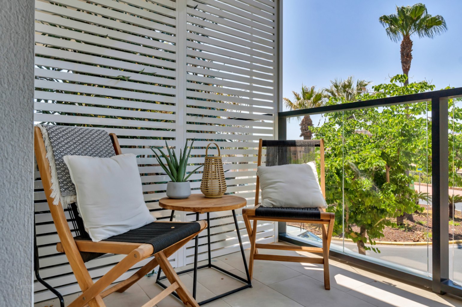 Privéterras met houten loungestoelen, een wit lamellenscherm en een glazen balustrade met uitzicht op de omliggende palmbomen en beplanting.