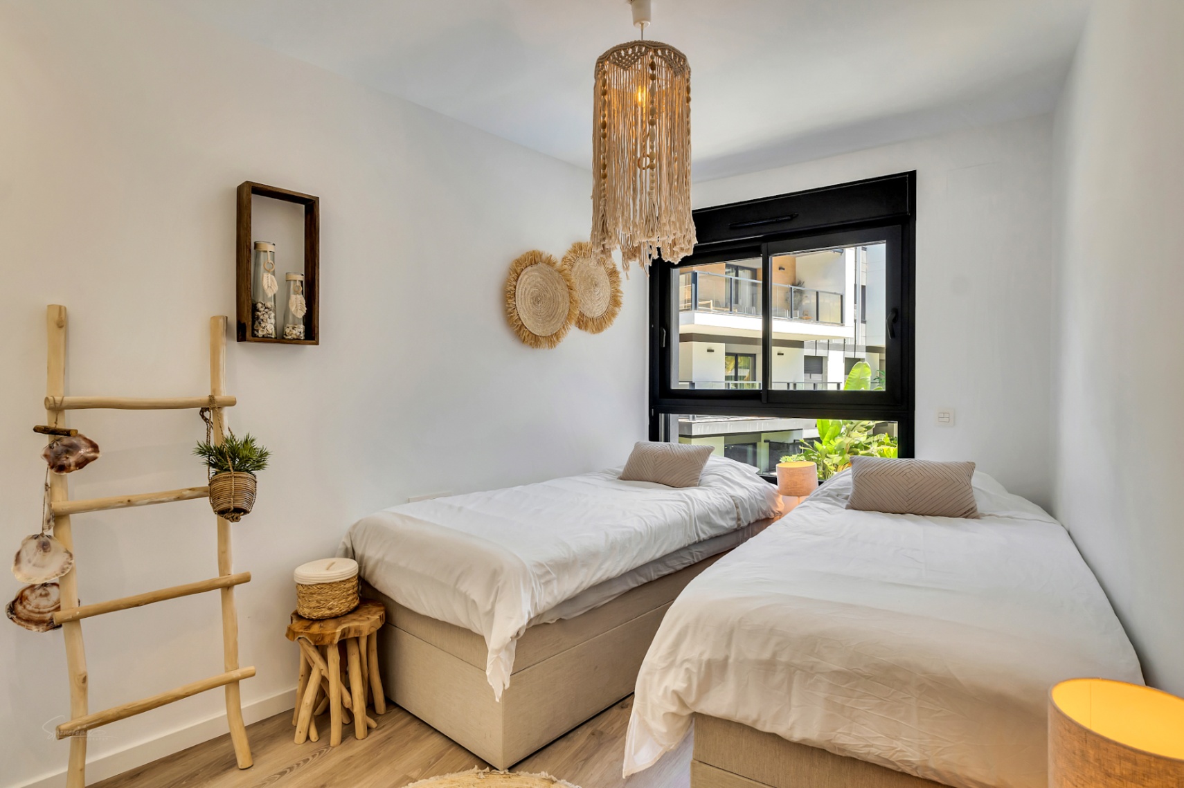 Slaapkamer met twee eenpersoonsbedden, mediterrane decoratie en een groot raam met zwart kozijn dat uitkijkt over de gemeenschappelijke ruimtes.