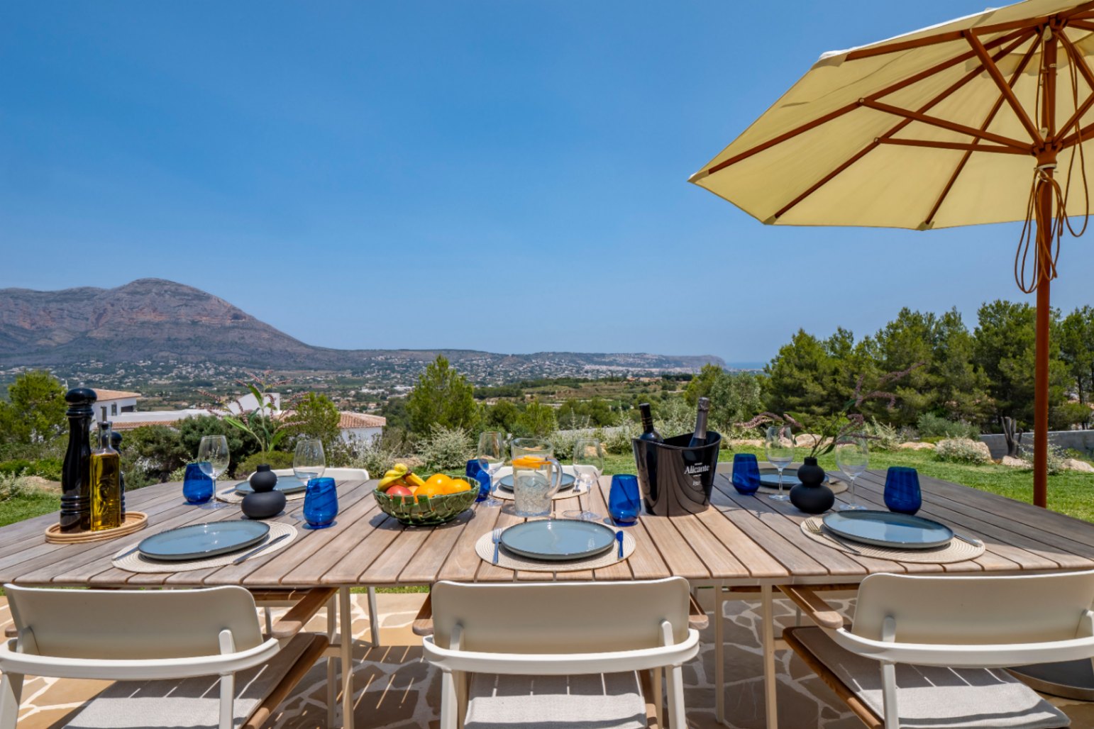 Eethoek buiten met houten tafel, blauw servies en panoramisch uitzicht op de bergen in Jávea.