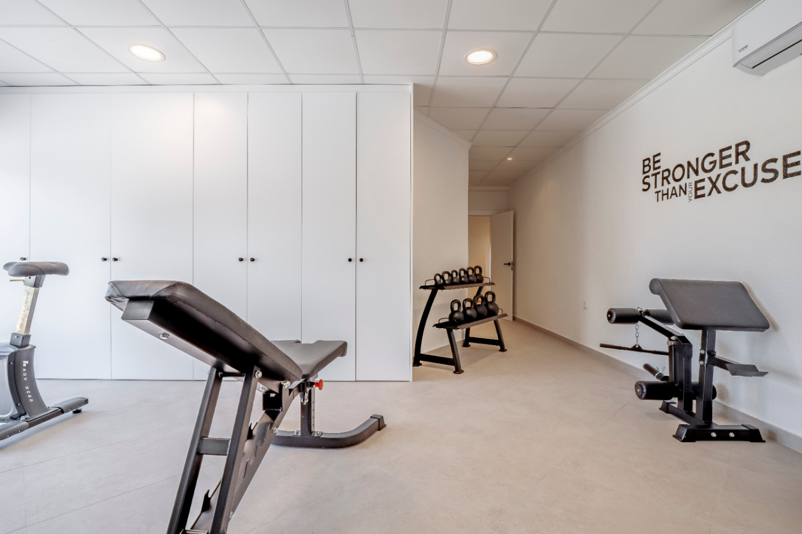 Fitnessruimte met keramische vloeren, witte muren en inbouwkasten. Trainingsapparatuur met gewichten en bank.