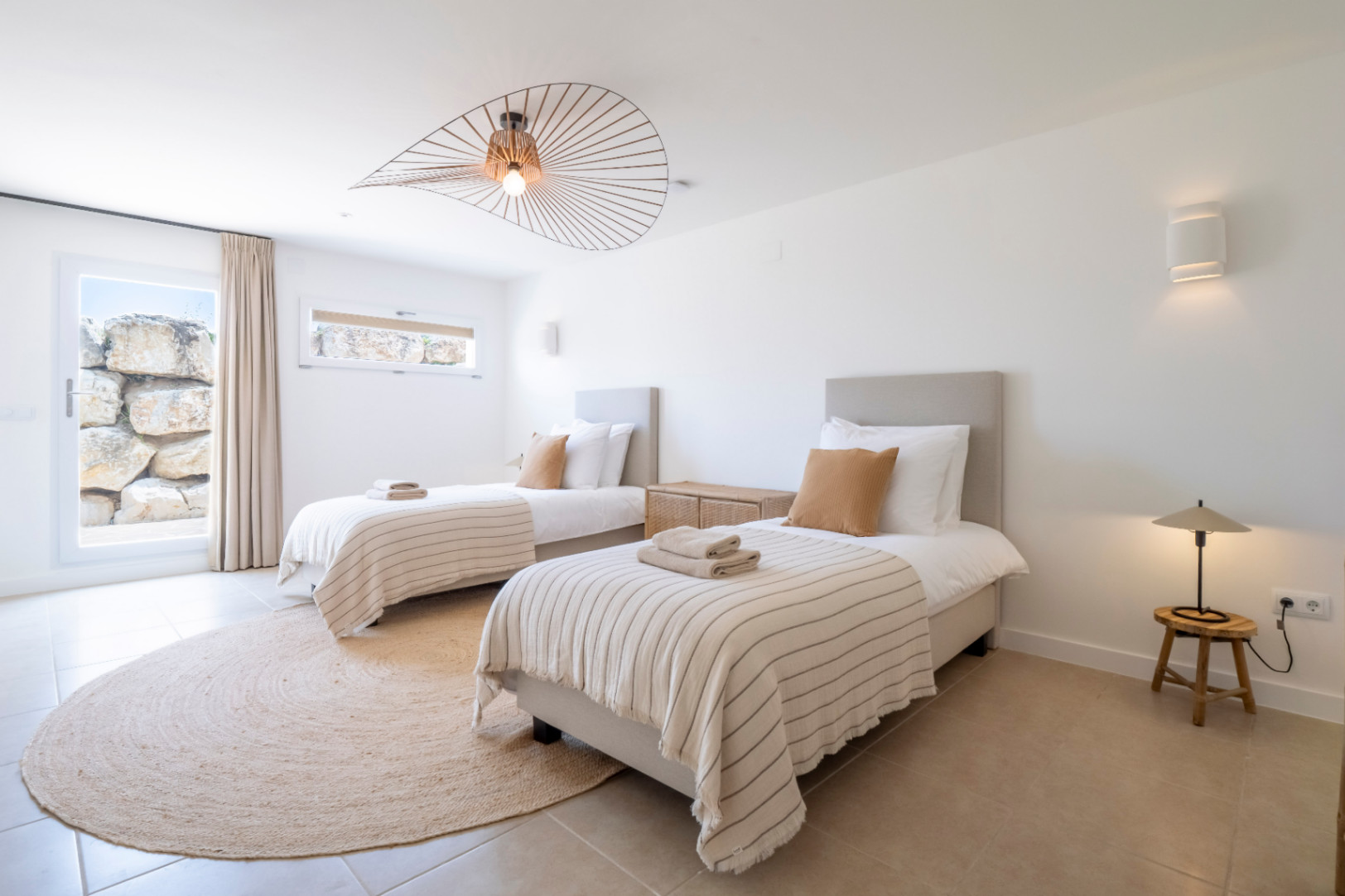 Lichte slaapkamer met twee eenpersoonsbedden, jute vloerkleed en designlamp.