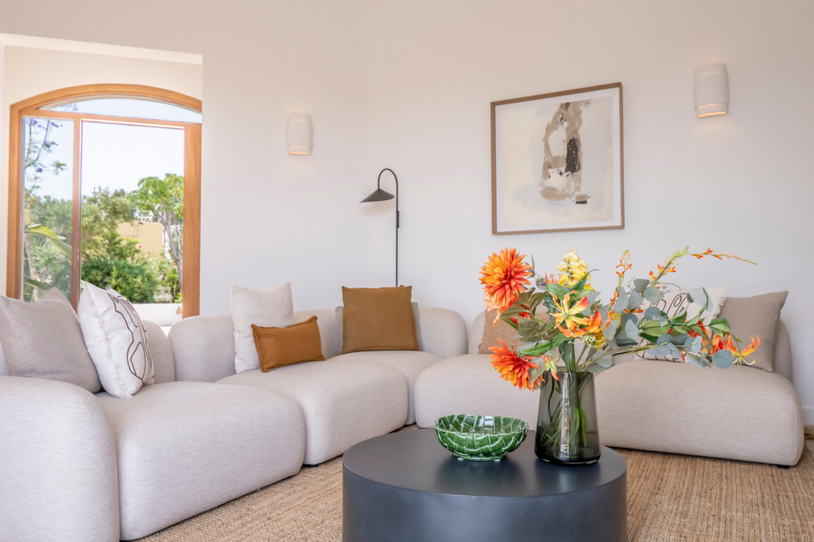 Lichte woonkamer met beige modulaire bank, bruine kussens en ronde zwarte tafel met bloemdecoratie.