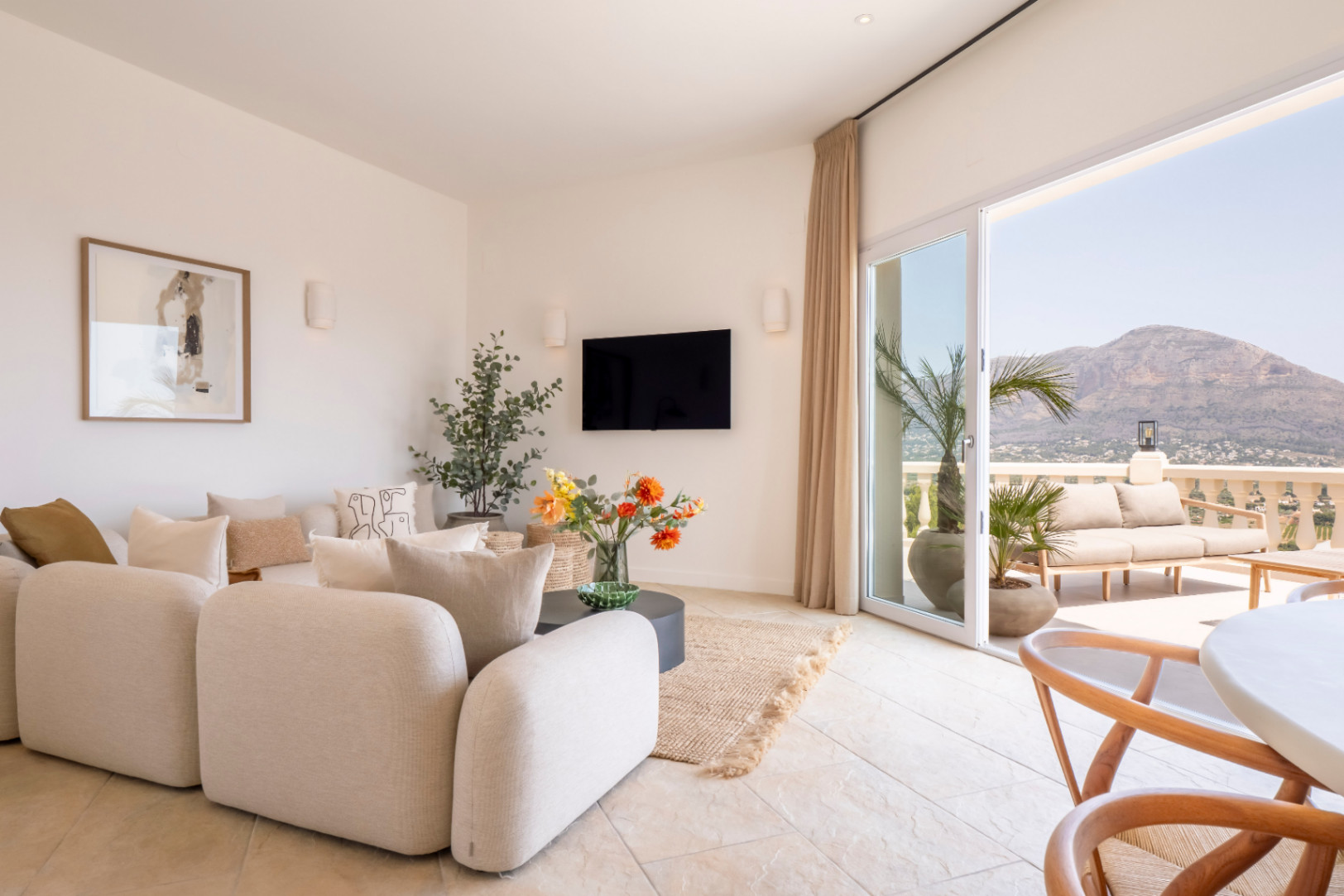 Lichte woonkamer met toegang tot terras. Beige modulaire bank, tv en bloemdecor. Berguitzicht in Jávea.