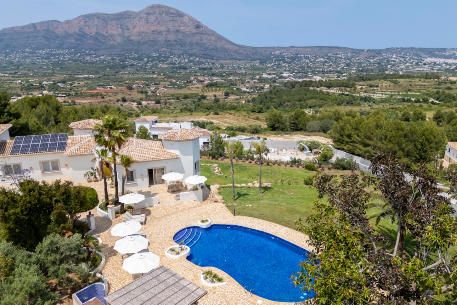 Luchtfoto van de villa met blauw zwembad, terrassen met witte parasols en een mediterrane tuin met uitzicht op de bergen.