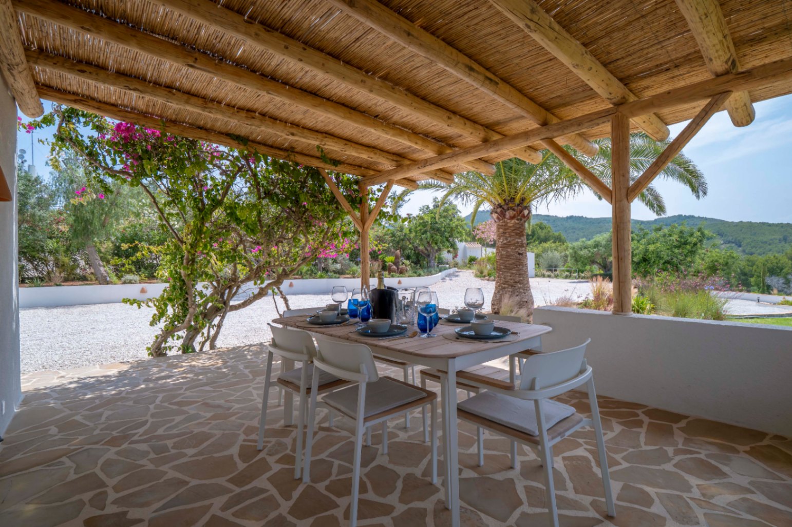 Overdekte eethoek buiten met rieten dak, onregelmatige stenen vloer en uitzicht op de mediterrane tuin.