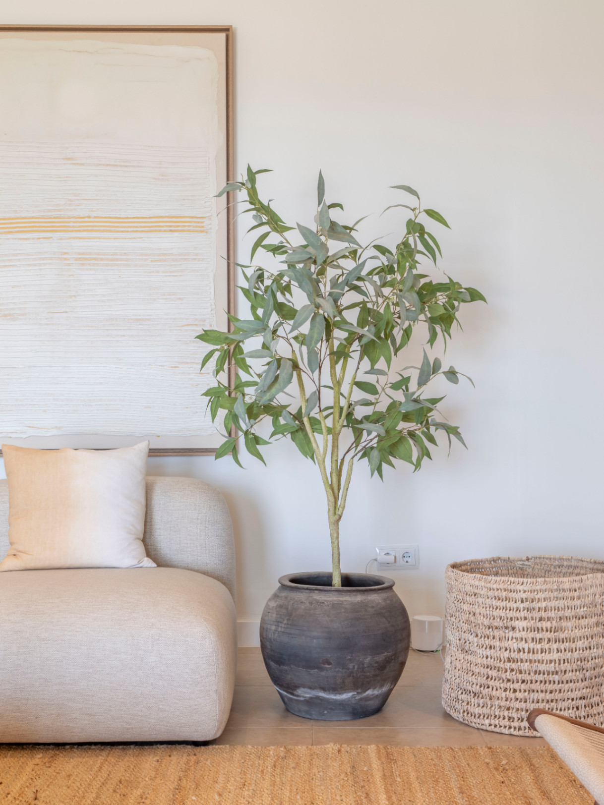 Woonkamerdetail met beige bank, keramische potplant en rieten mand op een jute vloerkleed.