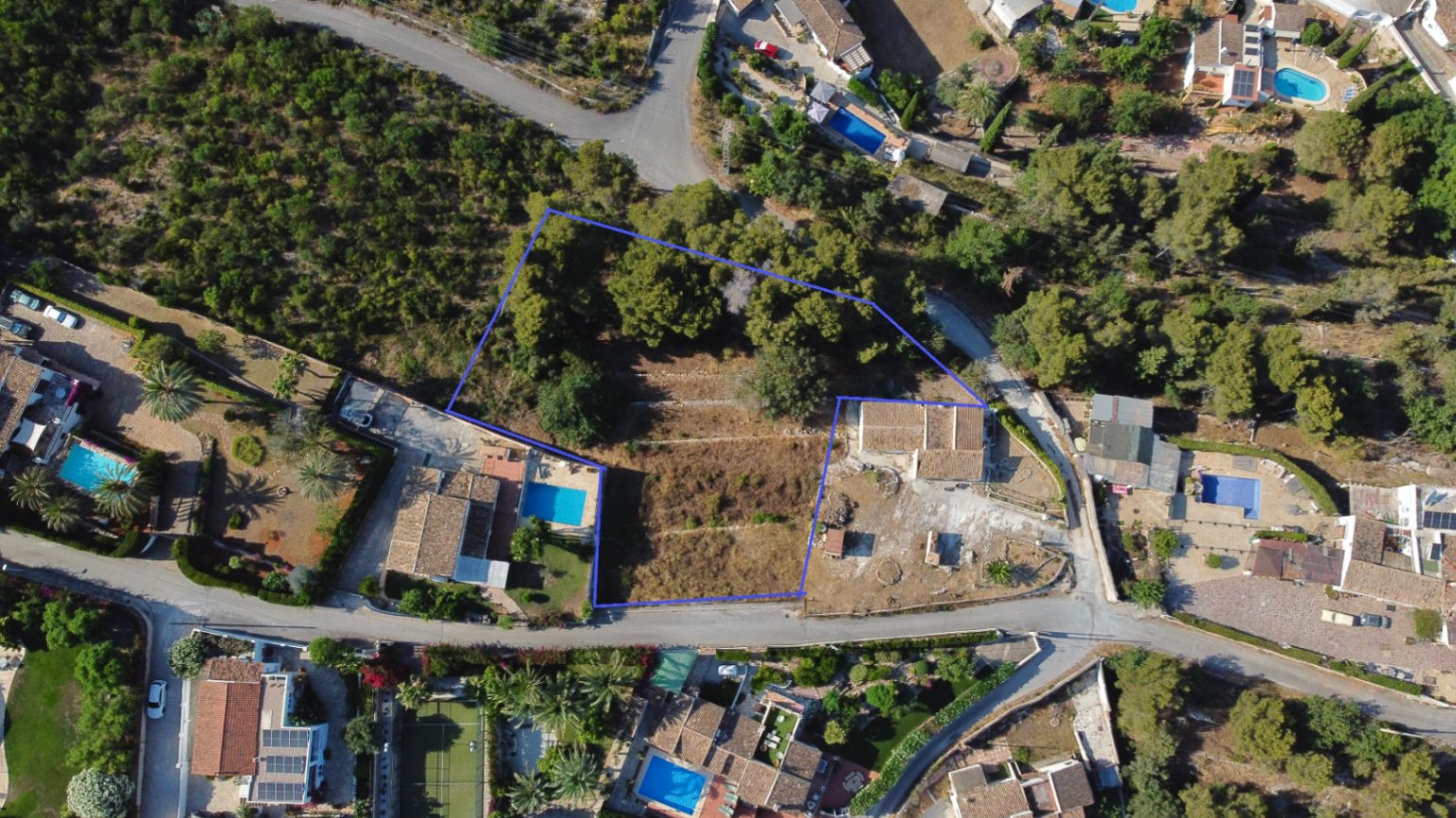Luchtfoto van het 1.908 m² grote perceel in Jávea met blauwe omlijning, terrasvormig terrein, volwassen pijnbomen en ligging nabij bestaande villa's en wegen.