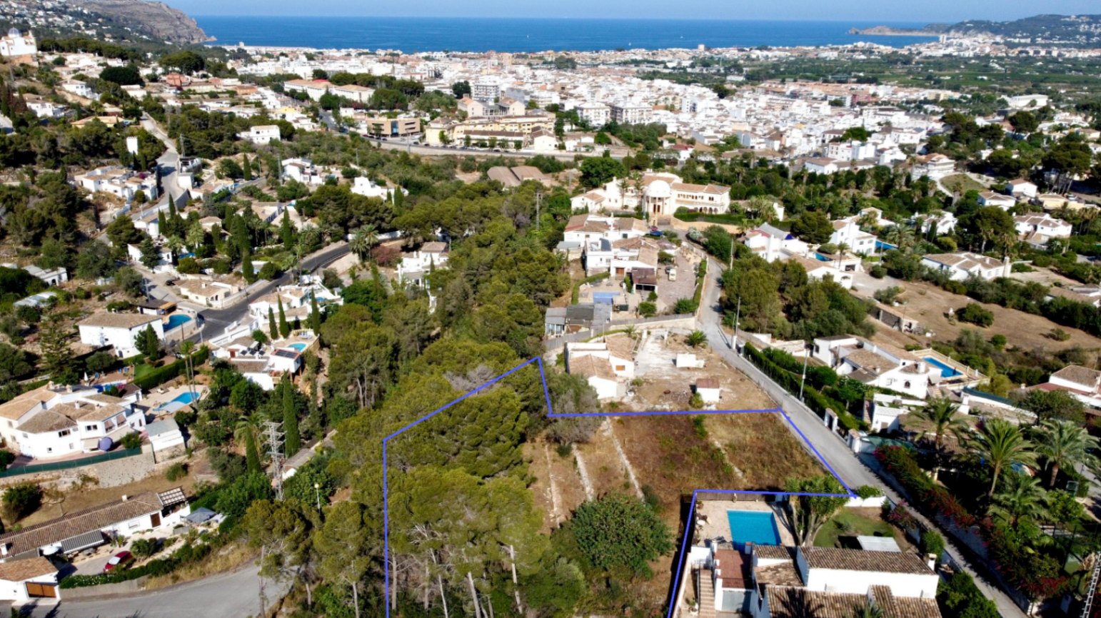 Luchtfoto van het blauw omlijnde perceel met pijnbomen, gelegen in een gevestigde woonwijk met uitzicht op de Middellandse Zee en het centrum van Jávea.