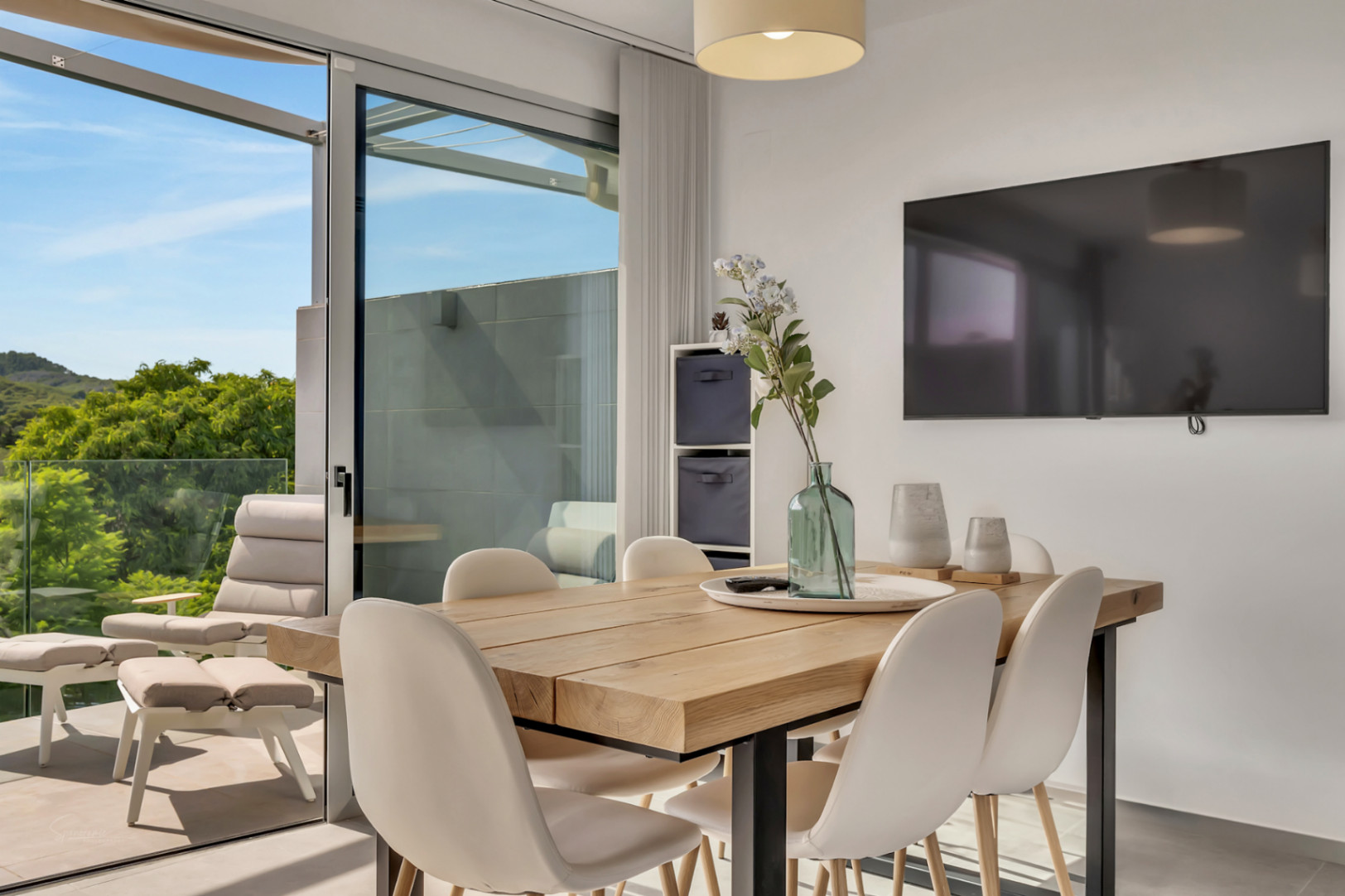 Eethoek met een massief houten tafel en witte stoelen, verbonden met een terras voorzien van loungemeubilair en uitzicht op de omliggende natuur.