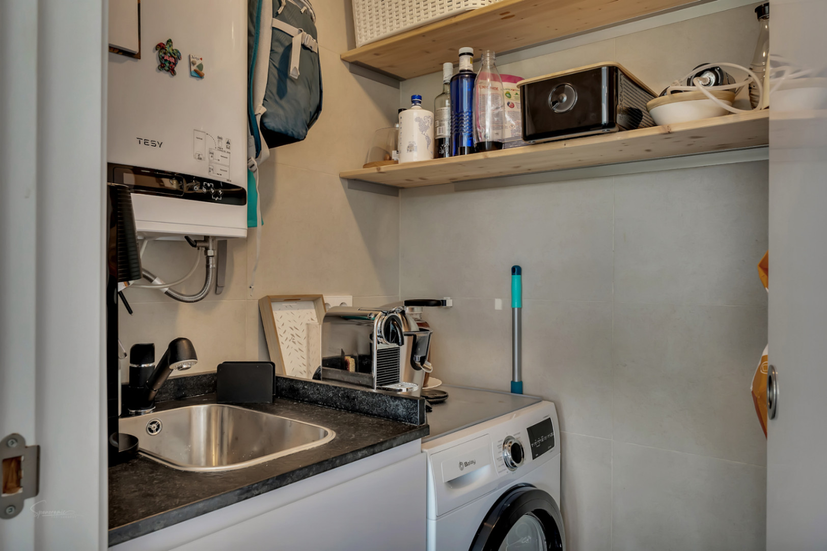 Functionele wasruimte uitgerust met een Balay wasmachine, roestvrijstalen spoelbak, Tesy elektrische boiler en houten planken.