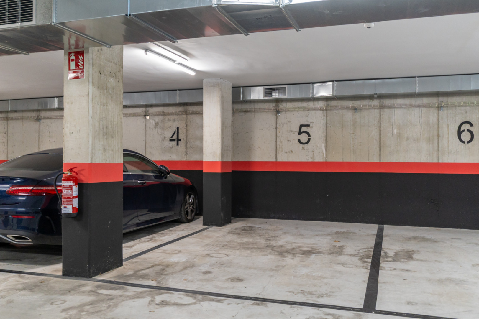 Genummerde ondergrondse parkeerplaats met wanden van zichtbeton, industriële ventilatiekanalen en aanwezige brandbeveiligingsvoorzieningen.