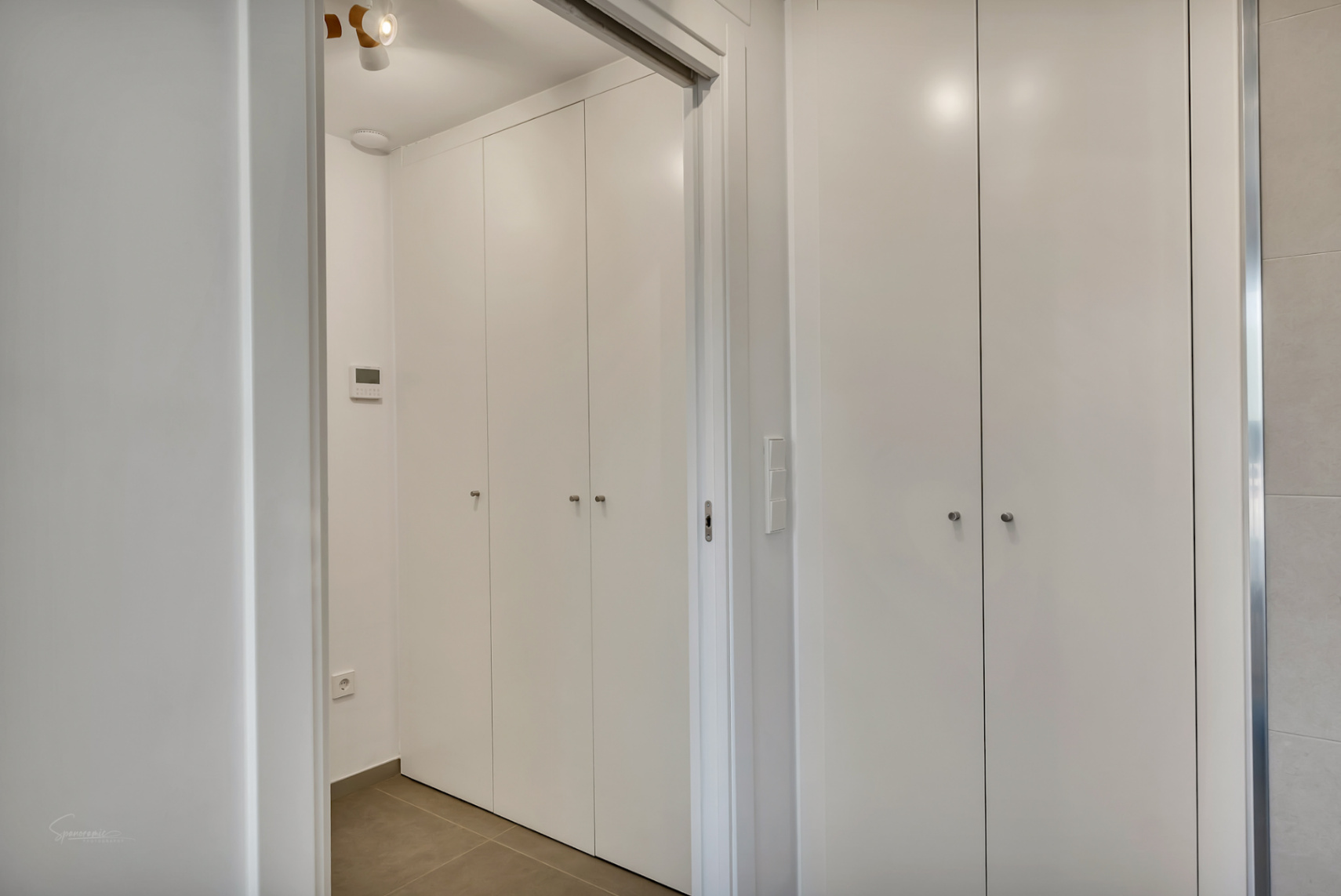 Kamerhoge inbouwkasten met een minimalistische witte afwerking en subtiele handgrepen, gelegen in een gang met een digitaal bedieningspaneel en moderne tegelvloer.