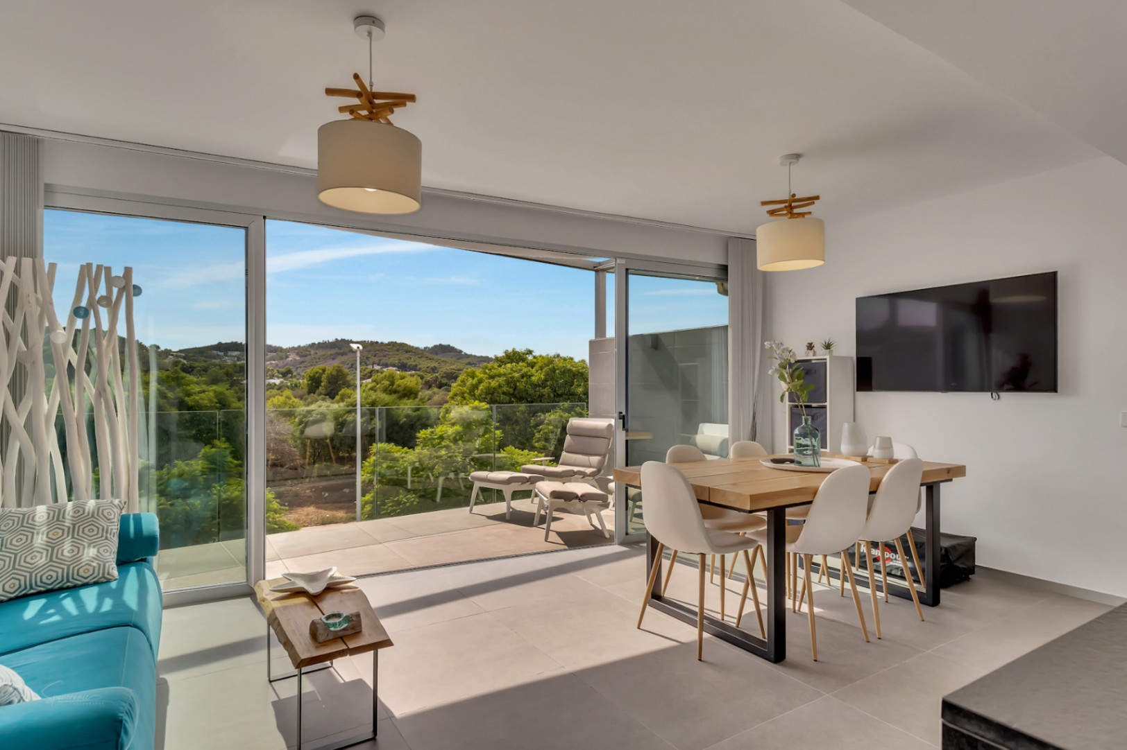 Lichte open woon- en eetkamer met grote glazen schuifdeuren naar een balkon met uitzicht op de natuur. Voorzien van een houten eettafel en moderne minimalistische inrichting.