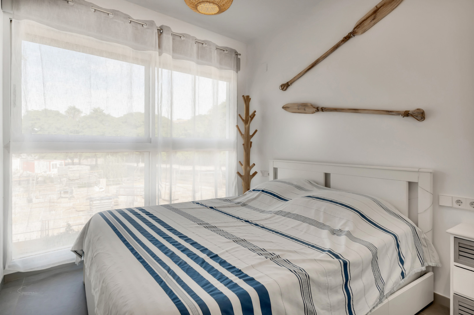 Lichte slaapkamer in nautische stijl met decoratieve houten roeispanen, een groot raam met veel lichtinval en een modern wit bedframe.