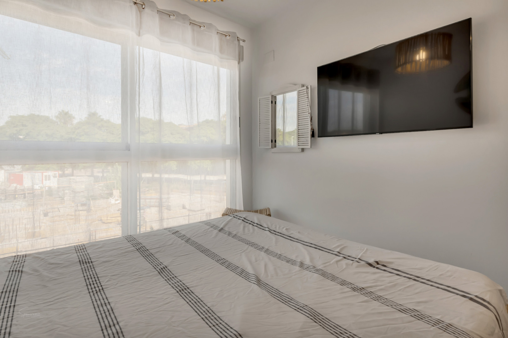 Lichte slaapkamer met een groot raam, doorschijnende gordijnen, een tweepersoonsbed met gestreept beddengoed en een aan de muur gemonteerde tv.