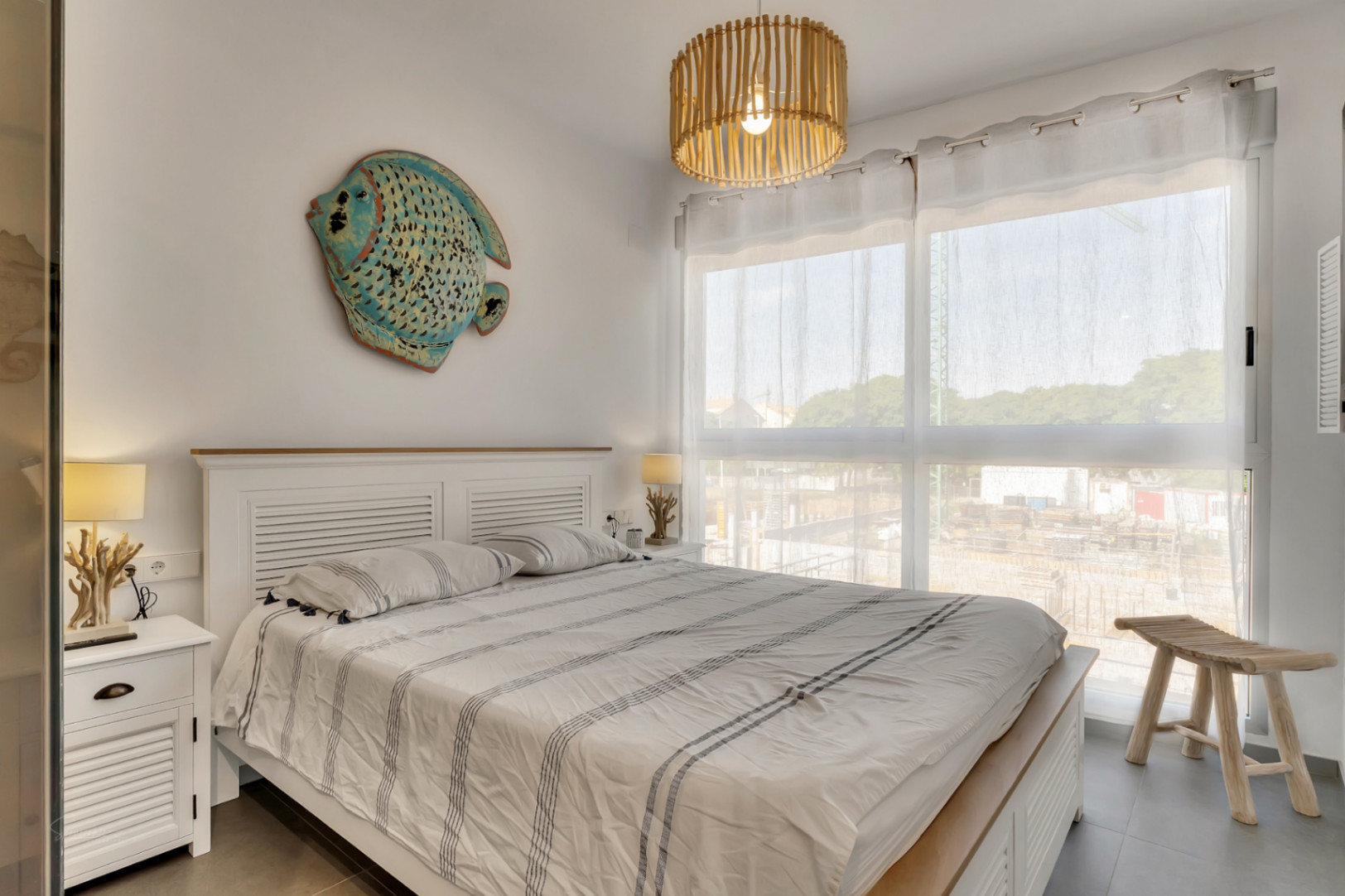 Lichte slaapkamer met wit houten bedframe, decoratie in maritieme stijl, een bamboe hanglamp en een groot raam voor natuurlijk licht.