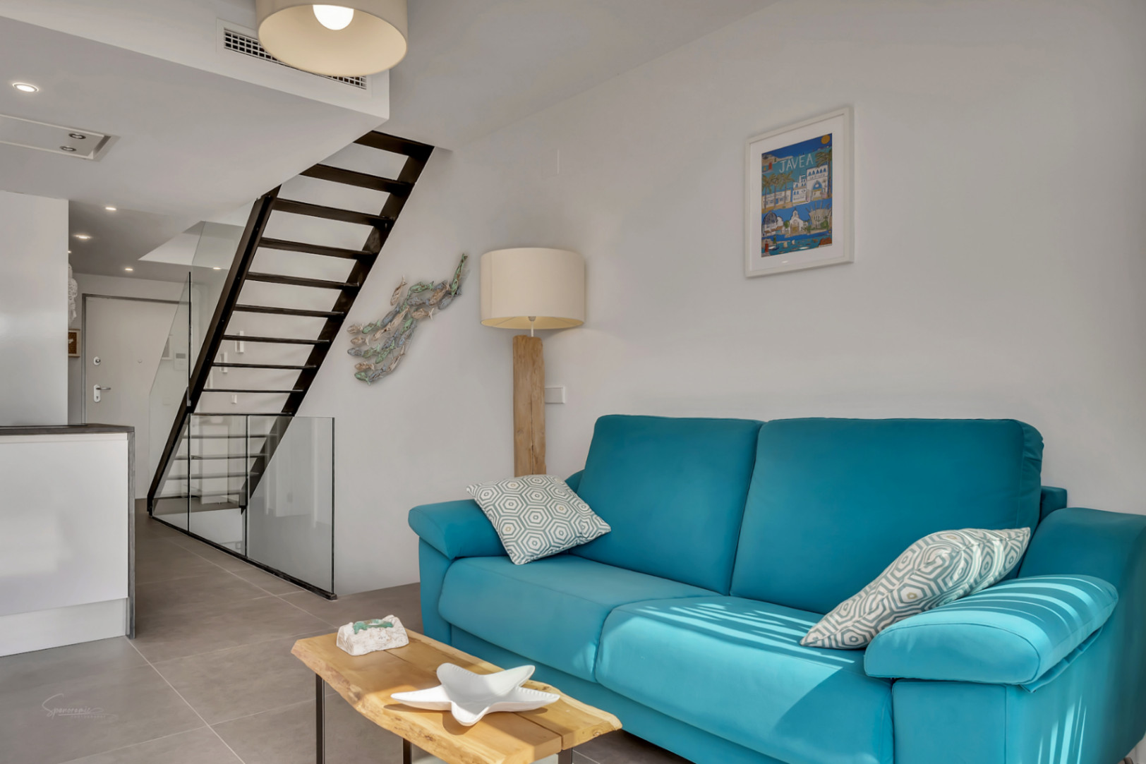 Lichte woonkamer met turquoise bank, natuurhouten tafel en een minimalistische zwarte metalen trap met glas. Grijze porseleinen tegelvloer en modern open ontwerp.