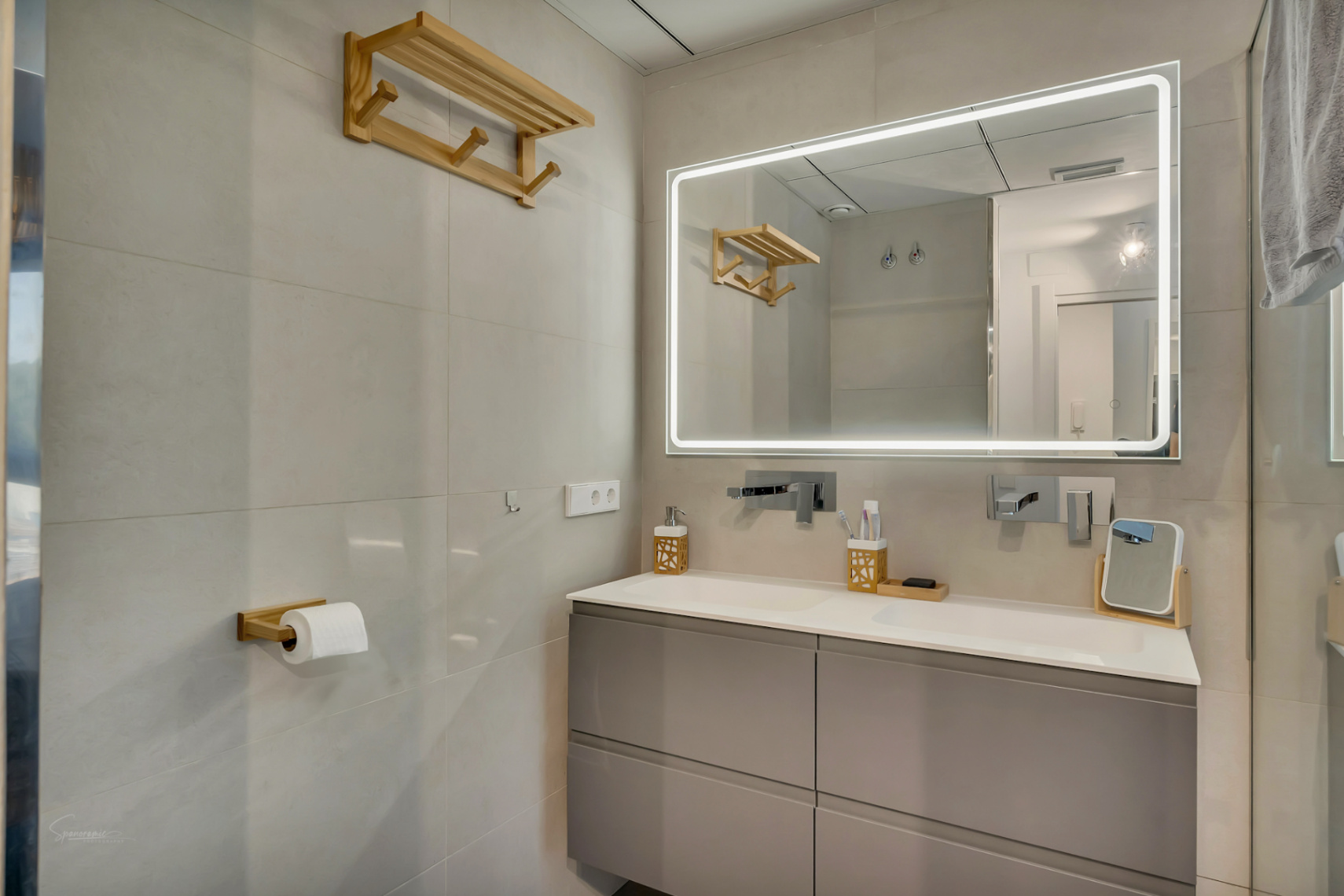 Moderne badkamer met dubbele wastafel, grijs meubel en een grote spiegel met LED-verlichting. Houten accenten en lichtgrijze wandtegels zorgen voor een strakke afwerking.