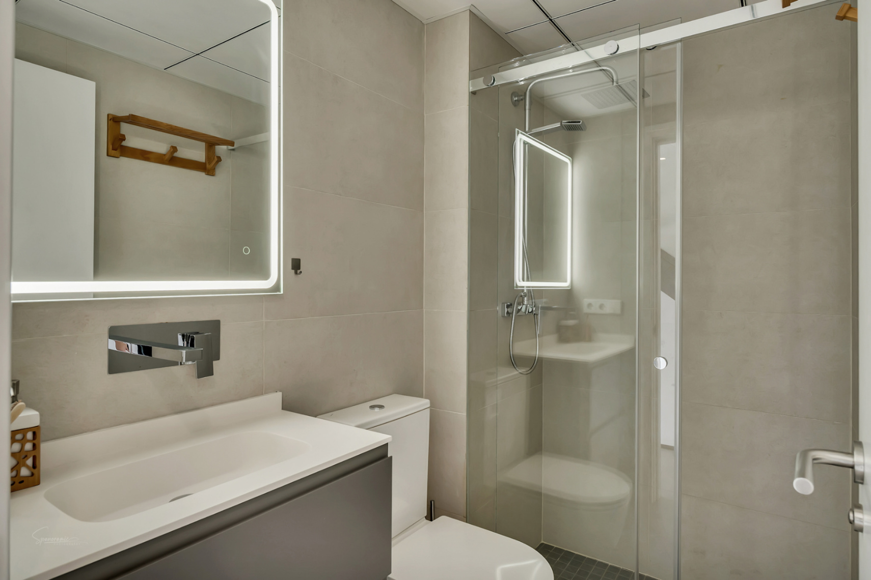 Moderne badkamer met neutrale tegels, een grijs meubel en een spiegel met LED-verlichting naast een inloopdouche met glazen schuifdeur.