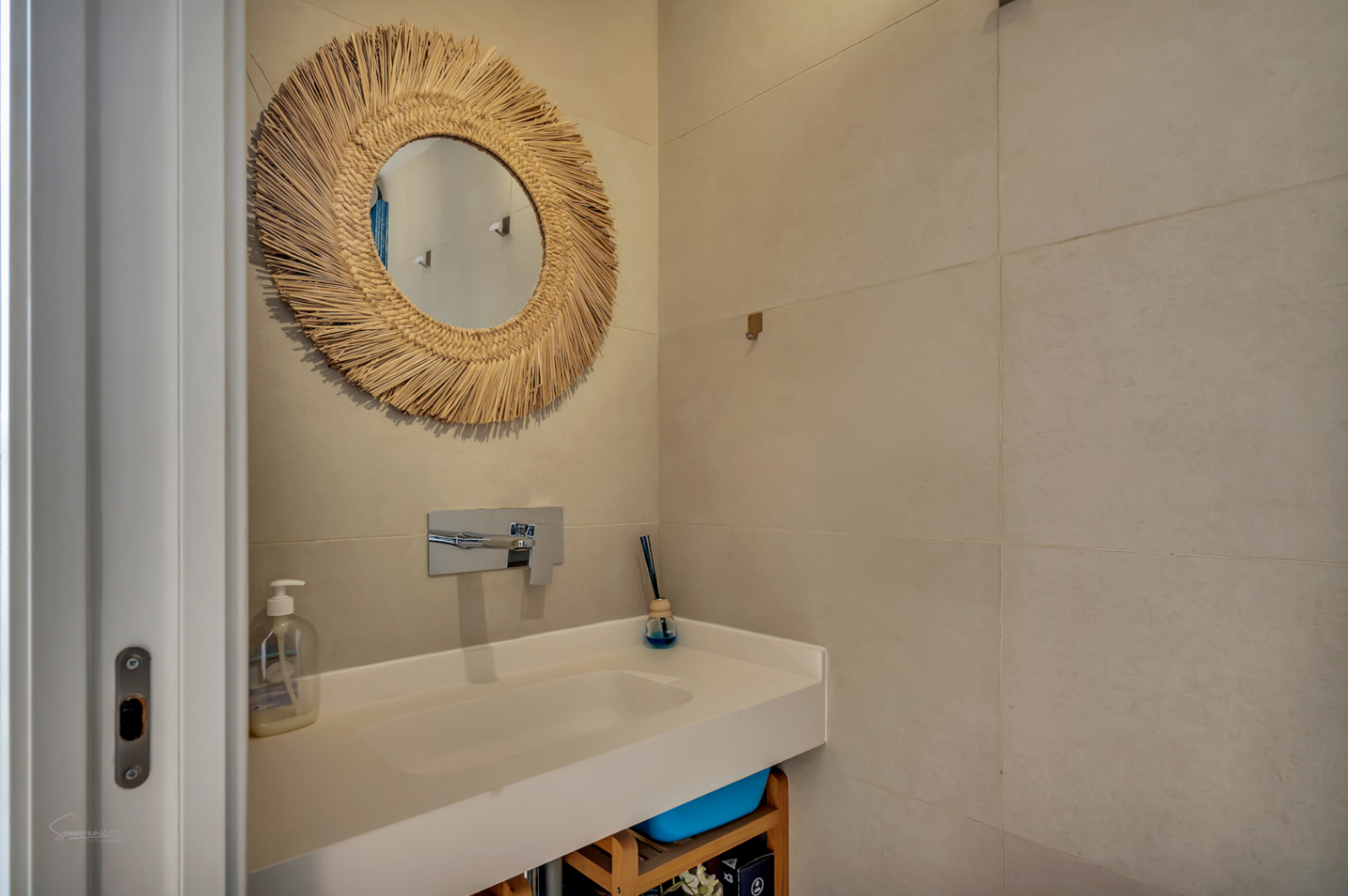 Moderne badkamer met wit geïntegreerd wastafelblad, chromen inbouwkraan en een decoratieve spiegel met natuurlijke vezellijst tegen zandkleurige grootformaat tegels.