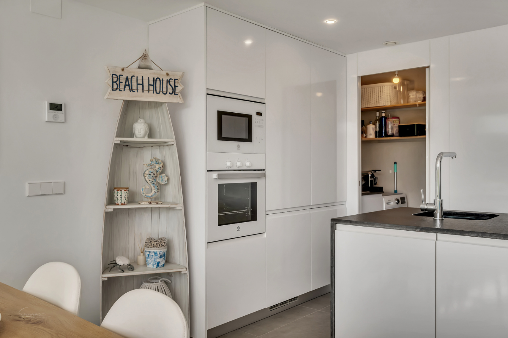 Moderne keuken met witte hoogglanskasten, geïntegreerde apparatuur en aangrenzende wasruimte met een decoratieve nautische stellingkast.