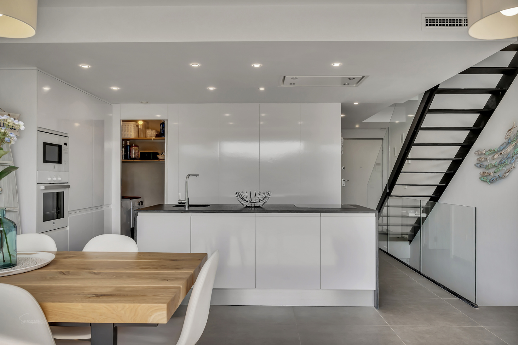Moderne minimalistische keuken met kookeiland, witte hoogglanskasten en een zwarte metalen trap met glas. De ruimte vloeit over in een eethoek met een massief houten tafel.