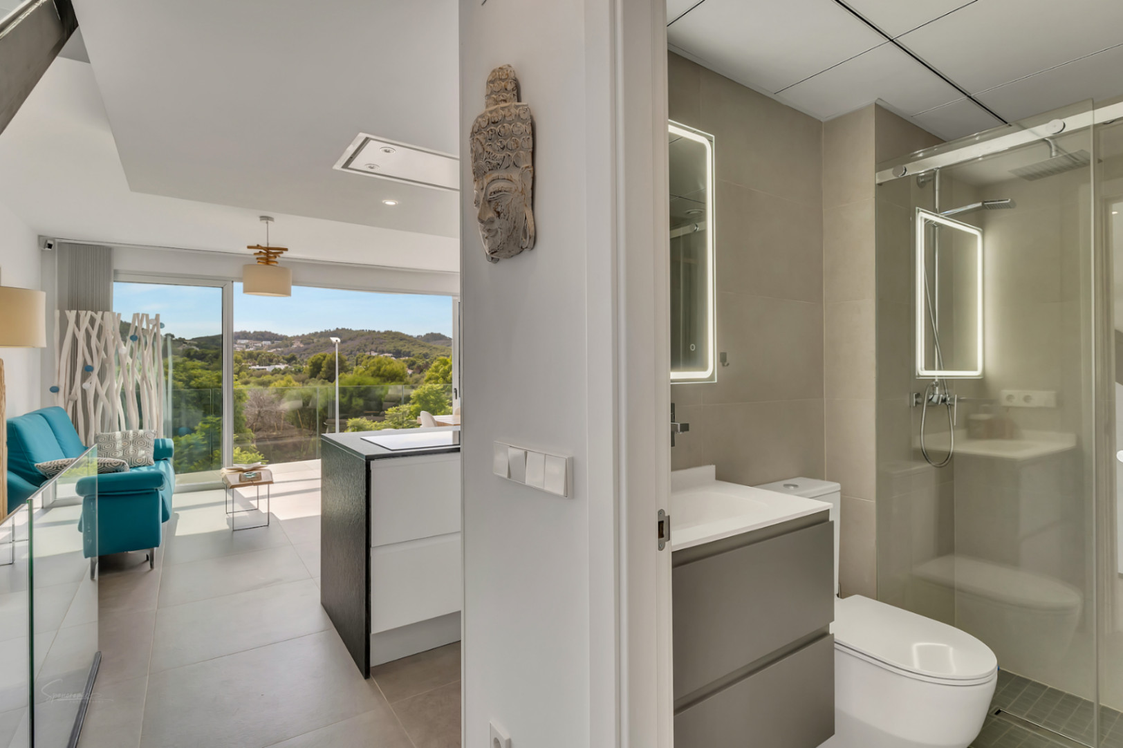 Zicht vanuit de gang op een moderne badkamer met verlichte spiegel en glazen douche, grenzend aan een lichte woonruimte met toegang tot het buitenterras.