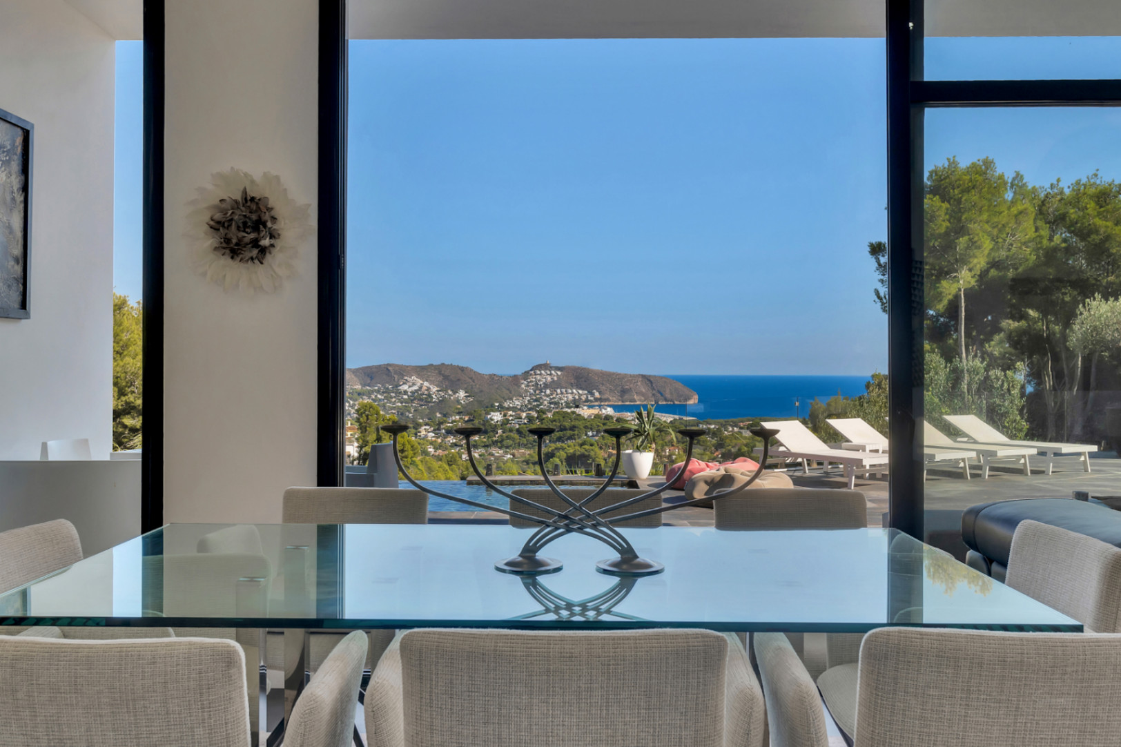 Eethoek met glazen tafel en gestoffeerde stoelen, met een panoramisch uitzicht op de Middellandse Zee door kamerhoge glazen schuifpuien.