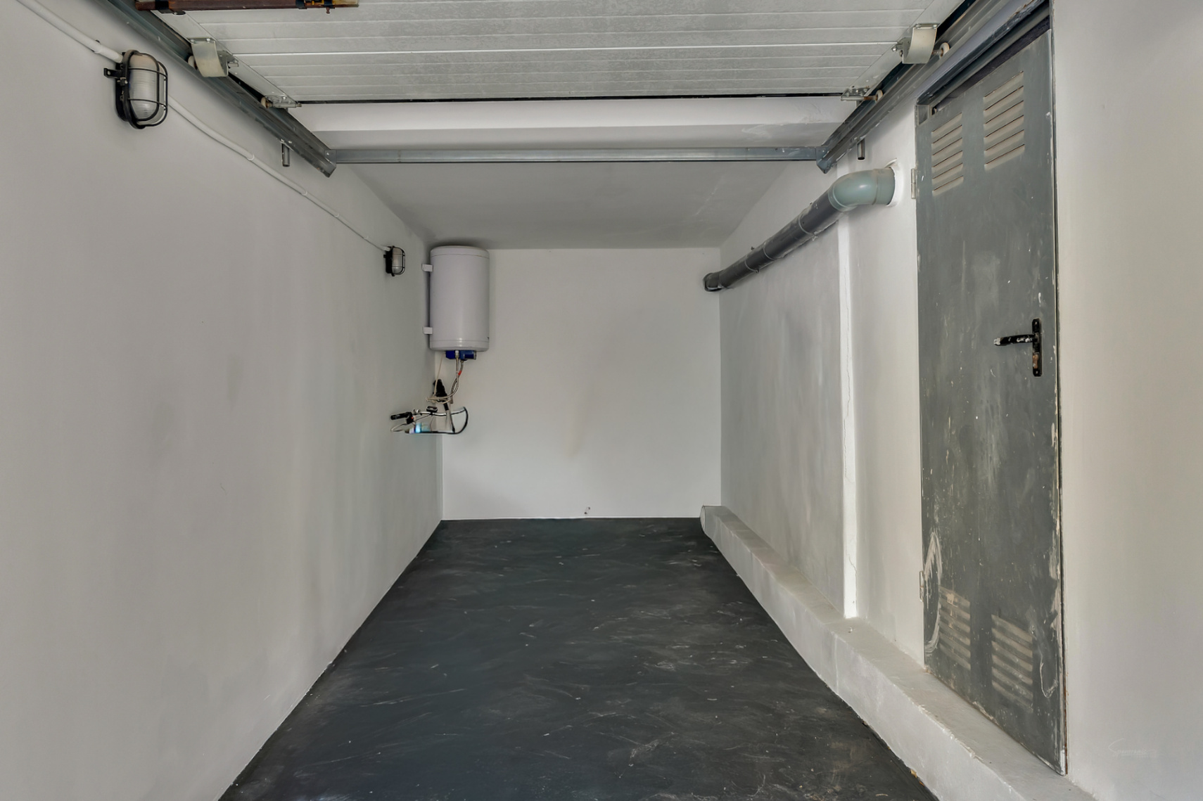 Functionele garage of bergruimte met donkere vloer, witte wanden, elektrische boiler en een metalen zijdeur met ventilatieroosters.
