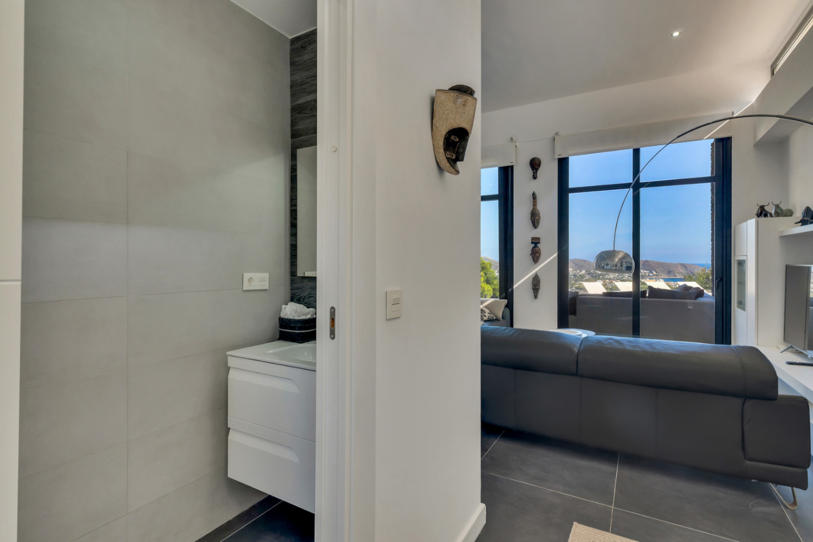 Gastentoilet met zwevend wit meubel grenzend aan de woonkamer, voorzien van grote raampartijen met uitzicht op zee en moderne decoratieve elementen.