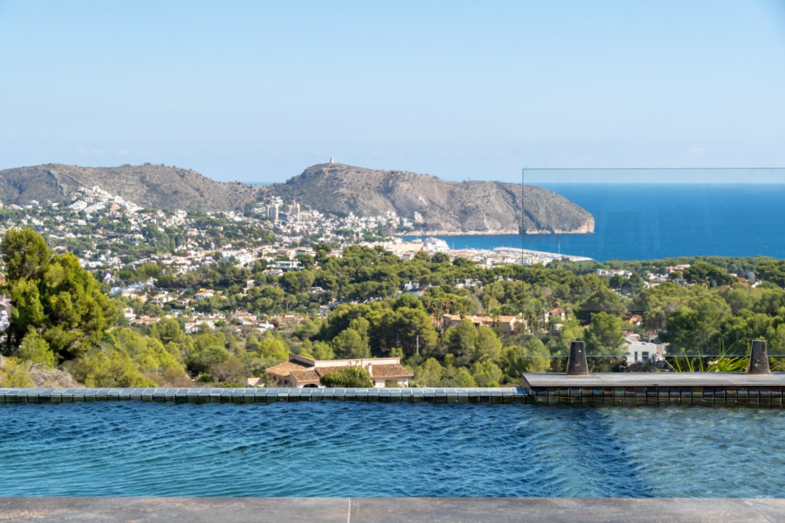 Infinity zwembad met blauwe mozaïektegels en glazen balustrade, met panoramisch uitzicht op de Middellandse Zee, Cap d'Or en de groene omgeving van Moraira.