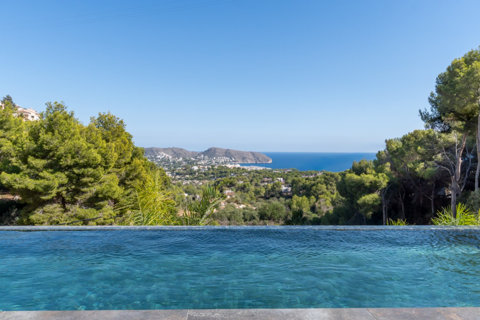 Infinity-zwembad met blauwe mozaïektegels en panoramisch uitzicht op de Middellandse Zee, de kustlijn van Moraira en het omliggende dennenbos.